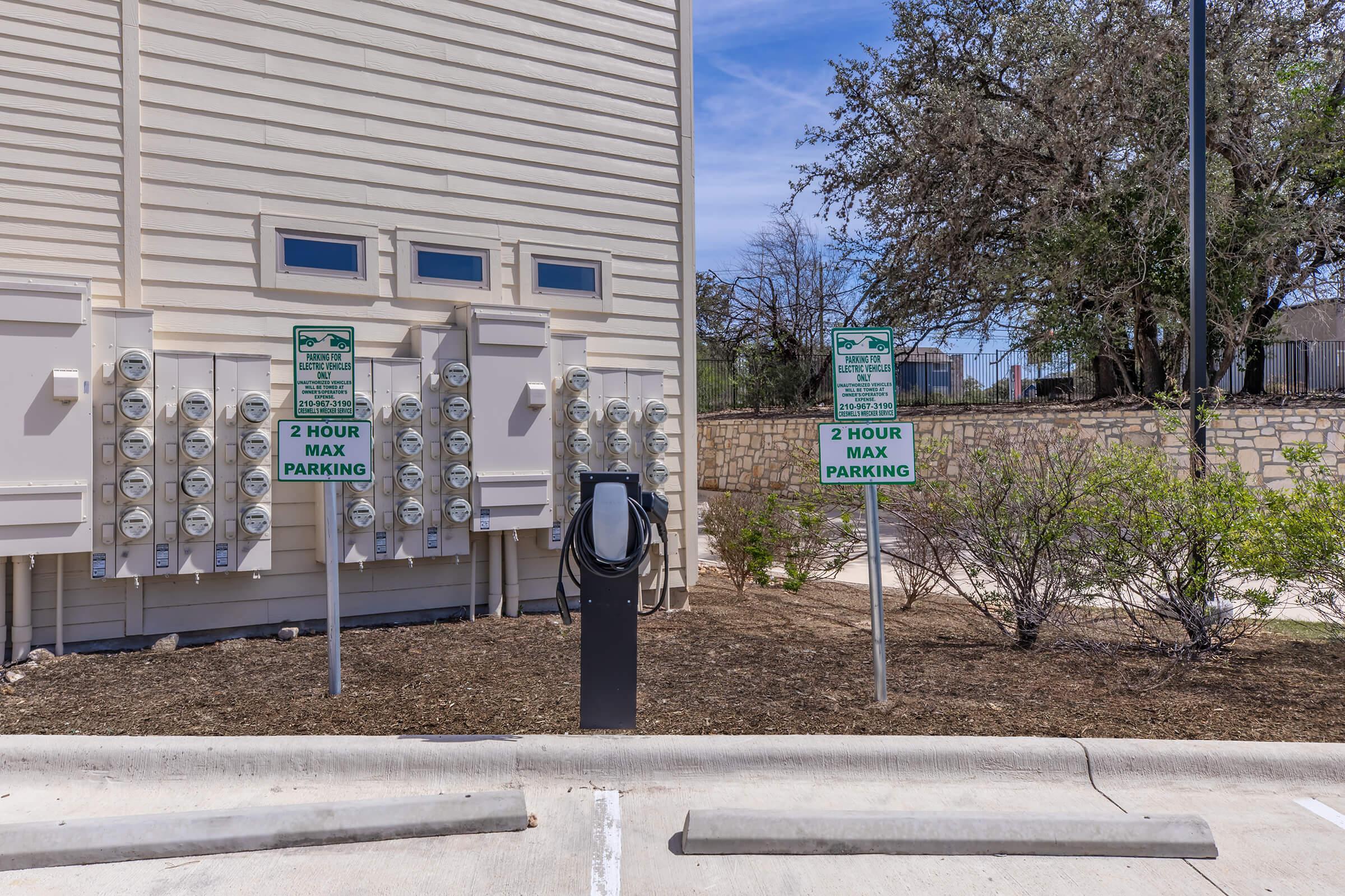 Electric vehicle charging station in a parking area, with multiple electric meters mounted on the building's wall. Signs indicate a maximum parking duration of 2 hours. Surrounding landscape includes low shrubs and a gravel area, with trees visible in the background.