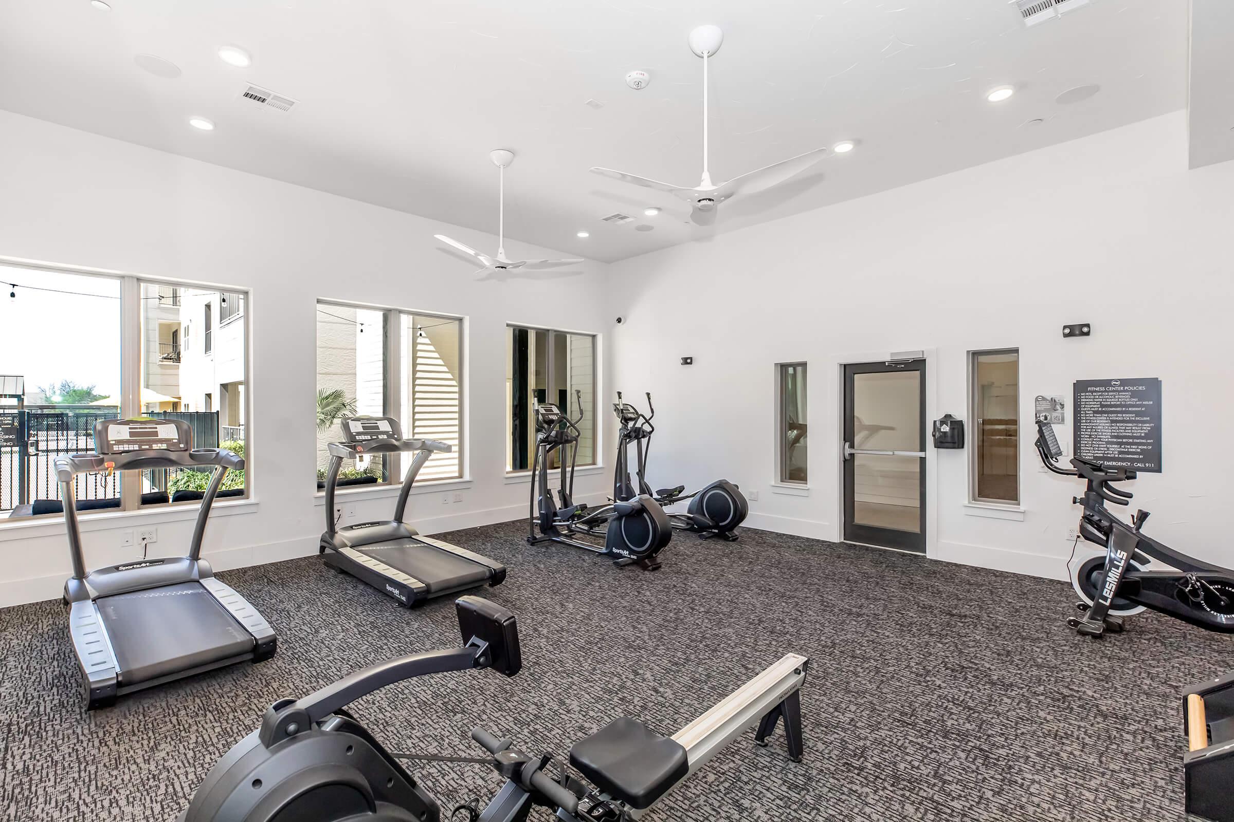 A modern gym interior featuring various workout equipment, including treadmills, ellipticals, and a rowing machine. Large windows provide natural light, and ceiling fans are installed for ventilation. The space has a clean, contemporary design with dark carpeting.