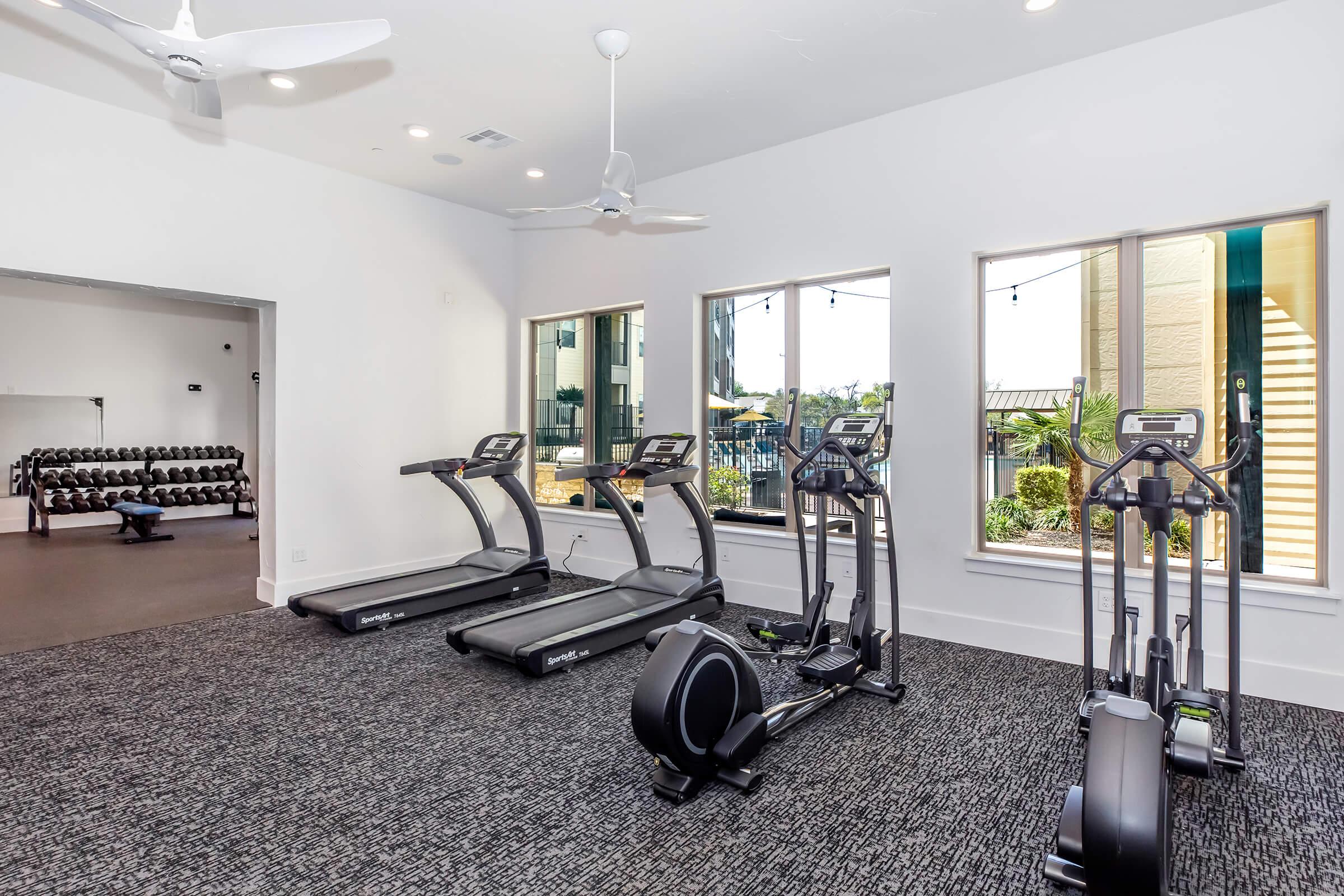 A modern gym featuring several cardio machines, including treadmills and an elliptical, with large windows allowing natural light. The space includes a weight area with dumbbells and a neatly carpeted floor. Ceiling fans are installed for air circulation, creating a welcoming workout environment.