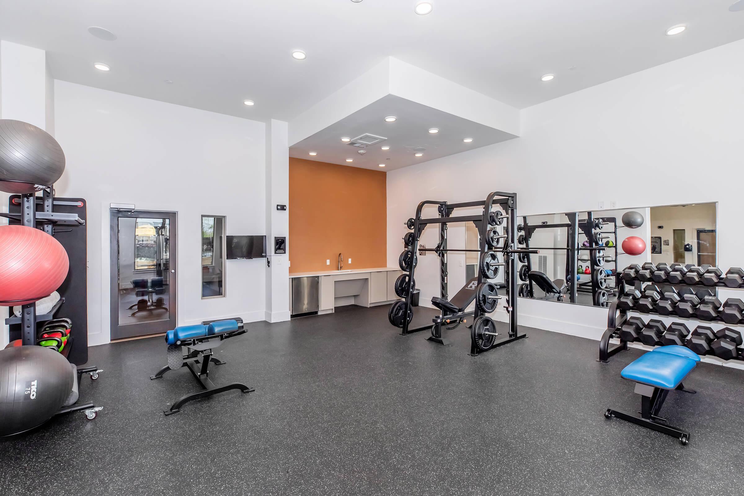 A modern fitness room featuring exercise equipment including weights, a bench, and stability balls. The space has a rubberized floor, mirrors on one wall, and bright overhead lighting, creating an inviting atmosphere for workouts.