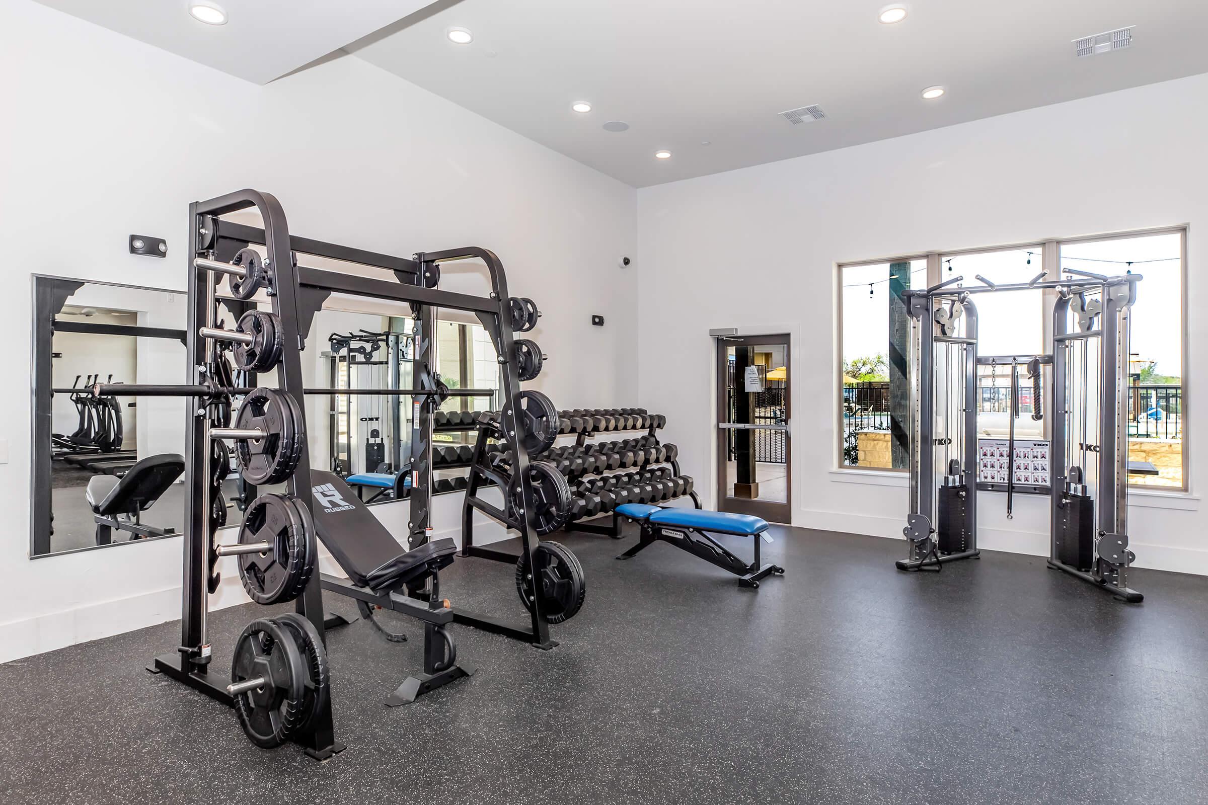 A modern gym interior featuring a squat rack with weights, a bench press, and various dumbbells on a rack. The flooring is dark, and there are large windows allowing natural light in. Mirrors are mounted on the walls, enhancing the open feel of the space. The environment is clean and well-maintained.