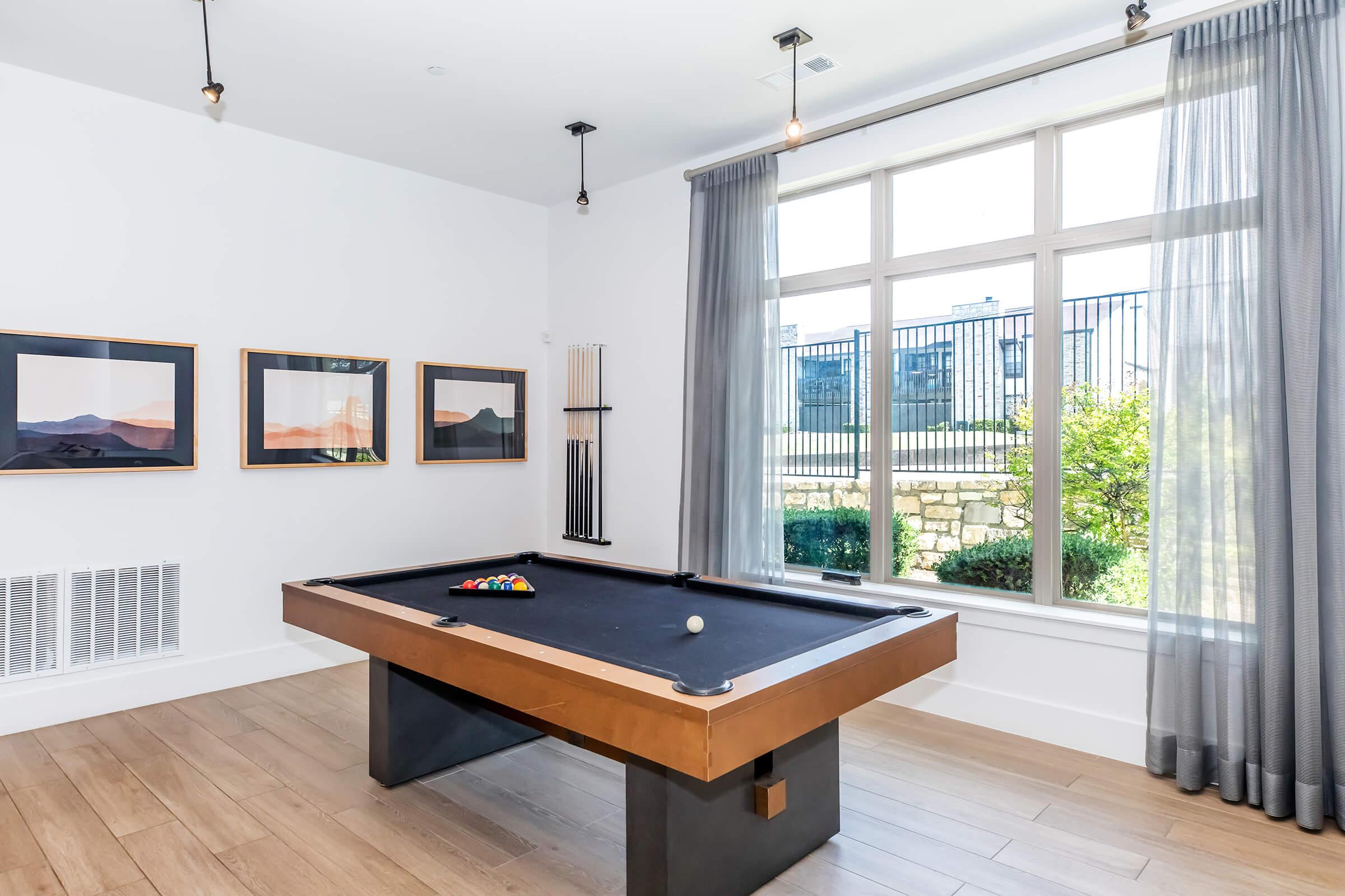A modern game room featuring a sleek black pool table with a single ball on it. The room has large windows draped with gray curtains, allowing natural light to illuminate the space. On the wall, there are framed landscape photographs, enhancing the contemporary aesthetic.