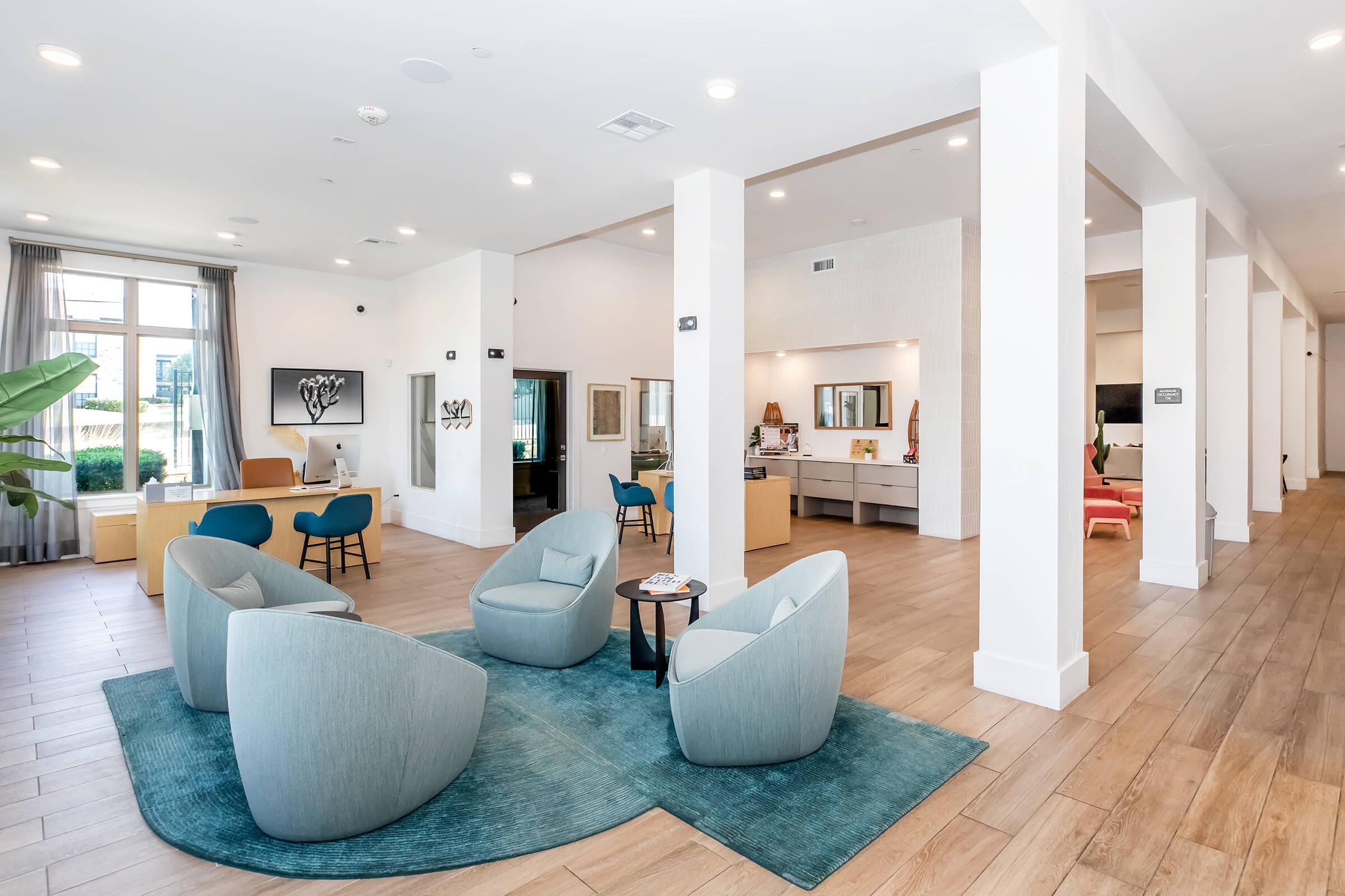 A modern lounge area featuring light-colored walls and hardwood flooring. Plush, rounded chairs in soft blue are grouped around a central table on a matching rug. A reception desk is visible in the background, along with additional seating areas that include colorful chairs and artwork on the walls. Natural light floods the space.