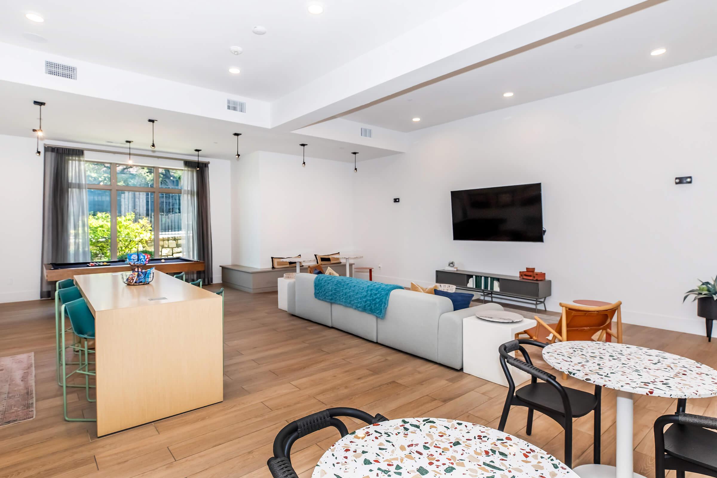 Modern living space featuring a light-filled room with a large television, a comfortable seating area with a gray sofa, and a dining table with circular patterned tops. There are green bar stools at a kitchen island and a cozy ambiance created by recessed lighting and natural light from large windows.