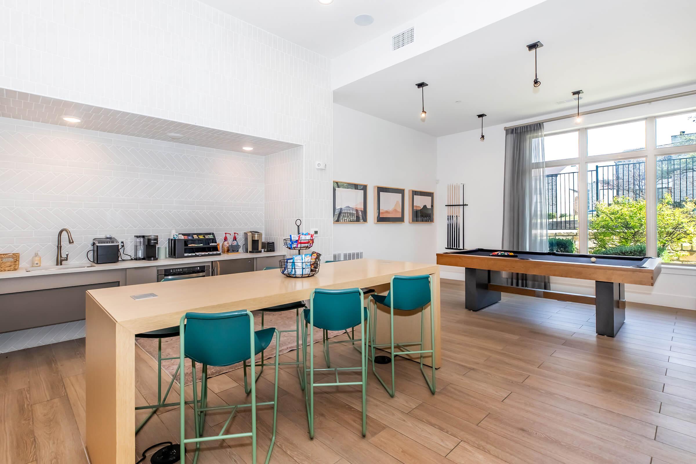 A modern kitchen and lounge area featuring a sleek countertop with seating, coffee machines, and decor. Large windows allow natural light, and there are framed pictures on the wall. A stylish pool table is positioned near a green outdoor view through the windows.