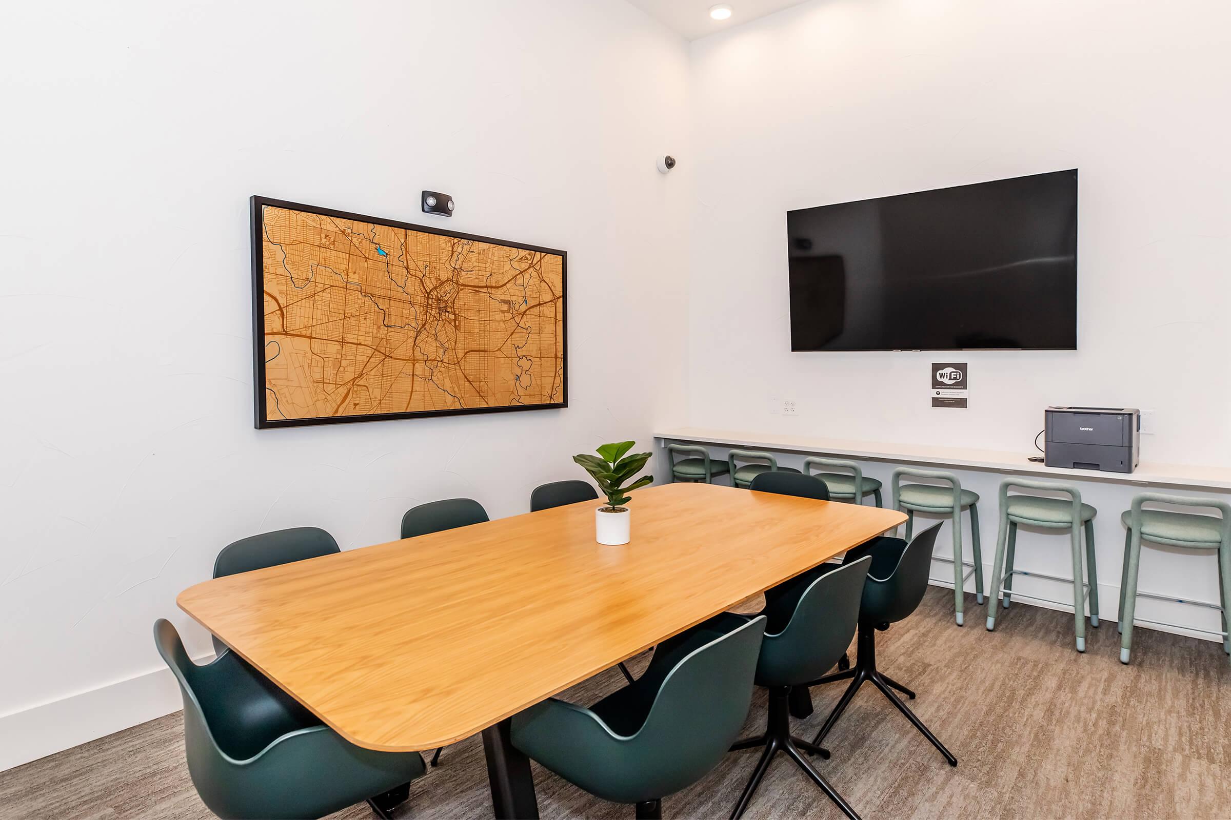A modern meeting room featuring a large wooden table surrounded by green chairs. On the wall hangs a framed map, and there is a large flat-screen TV mounted above a counter with bar stools. A small plant is placed in the center of the table, emphasizing a contemporary and inviting atmosphere.