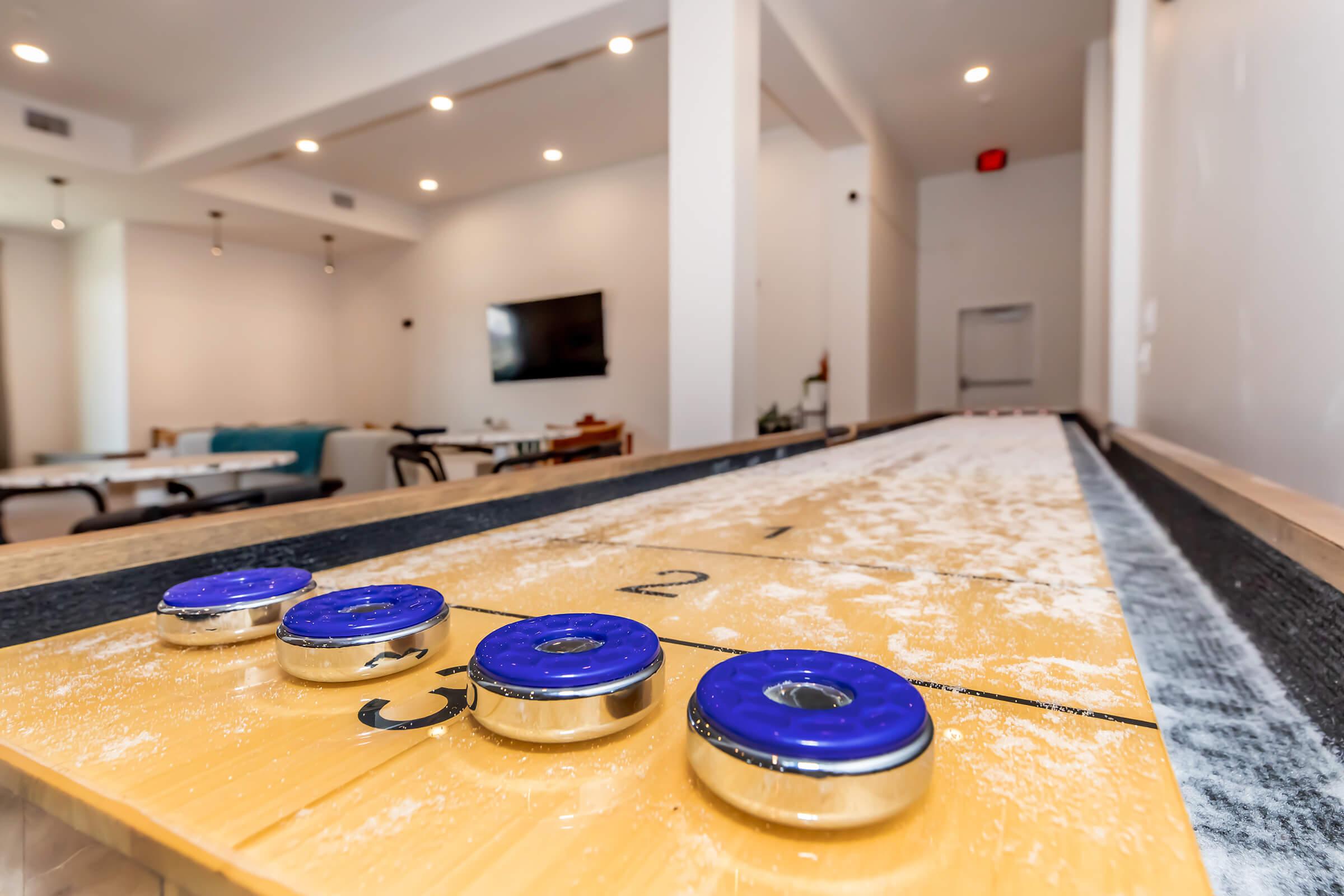 A close-up of a shuffleboard table with four blue pucks on it, positioned on a wooden surface marked with numbers. The background features a modern, bright room with minimalist decor, including a television and furniture, suggesting a recreational space for socializing and games.