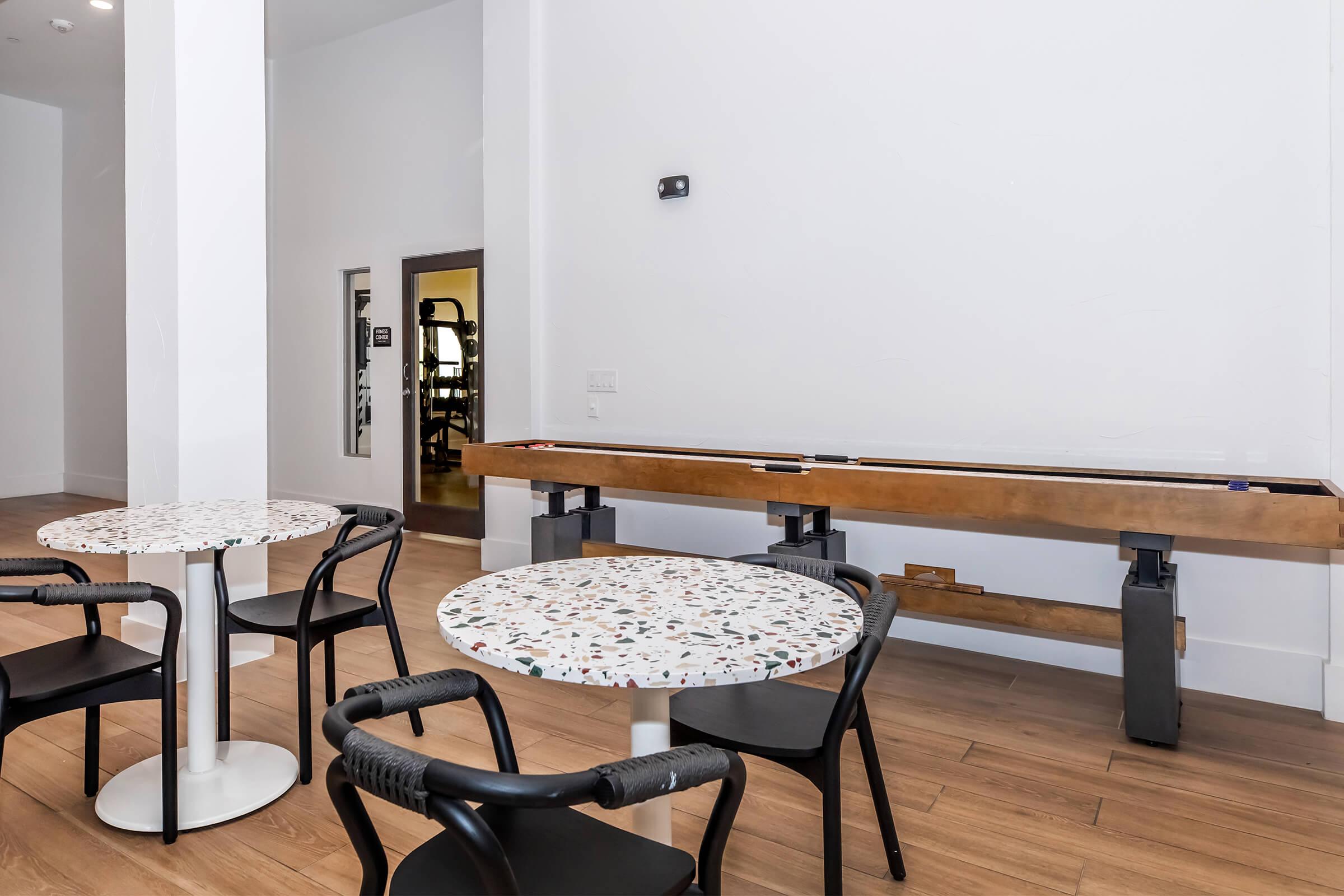 A spacious room featuring two round tables with patterned surfaces and black chairs. Along one wall, there is a wooden shuffleboard table. An open doorway leads to a fitness area at the back, with natural light illuminating the modern interior design.