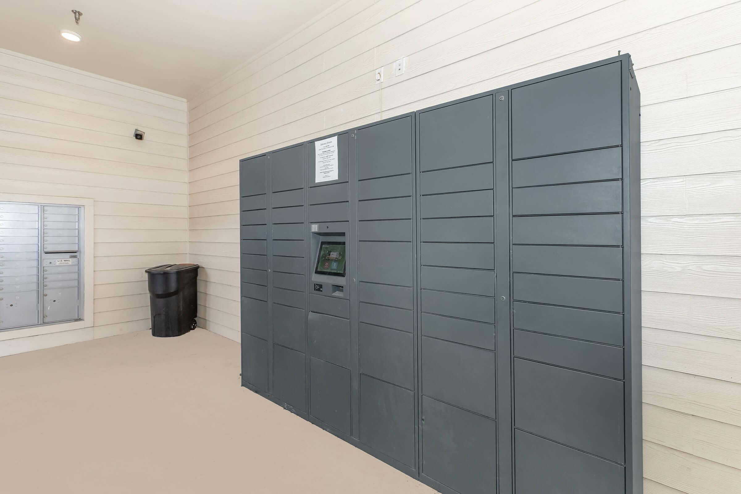 A large gray package delivery locker system installed against a light-colored wooden wall. There is a small screen and interface on the front of the locker. To the left, a standard mailbox can be seen, and a black trash bin is positioned against the wall in the background. The floor is light and smooth.