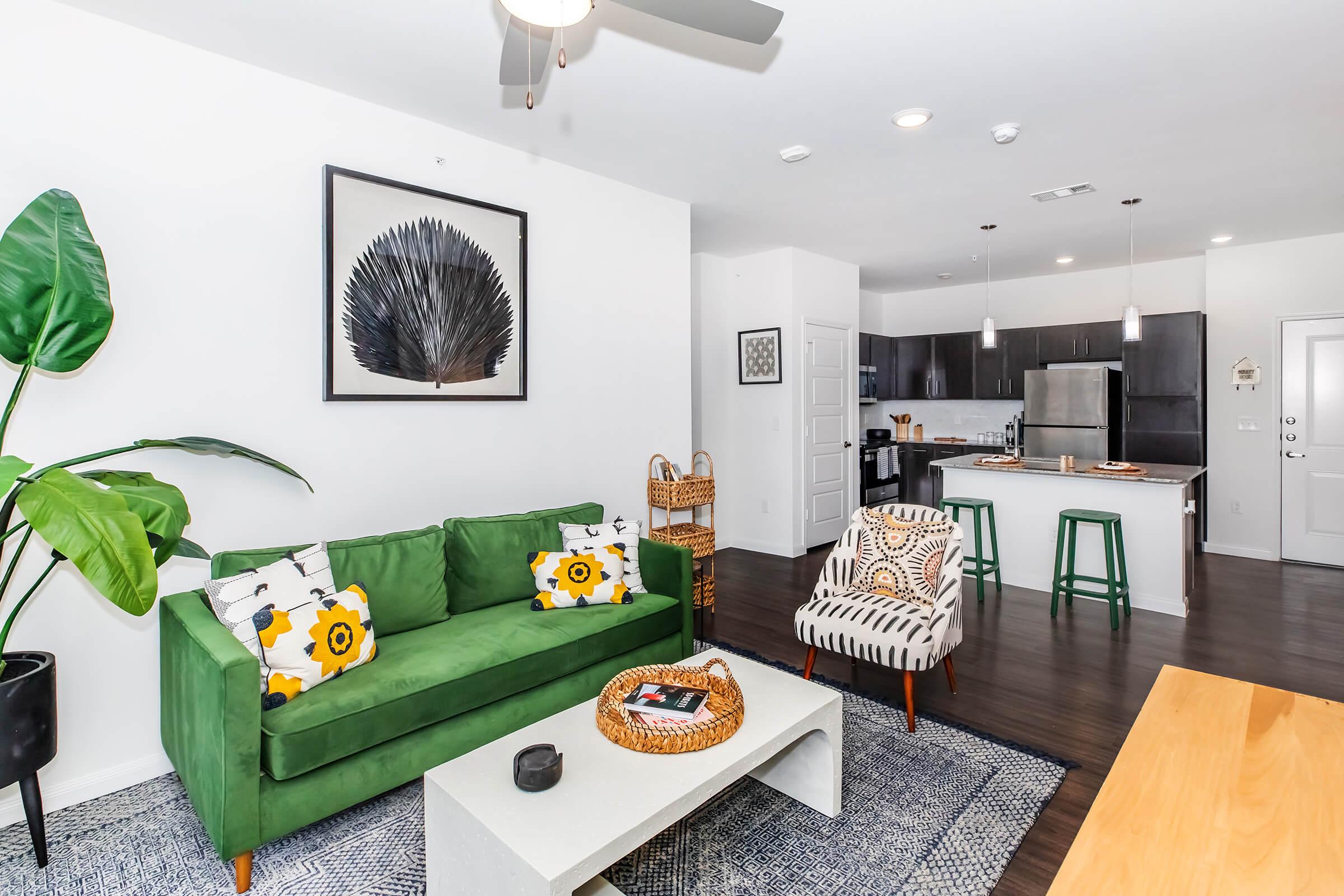 A modern living room featuring a green sofa with floral cushions, a patterned armchair, and a coffee table with decorative items. A large leafy plant stands in the corner, and the open kitchen with dark cabinetry and stainless appliances is visible in the background. Natural light brightens the space.