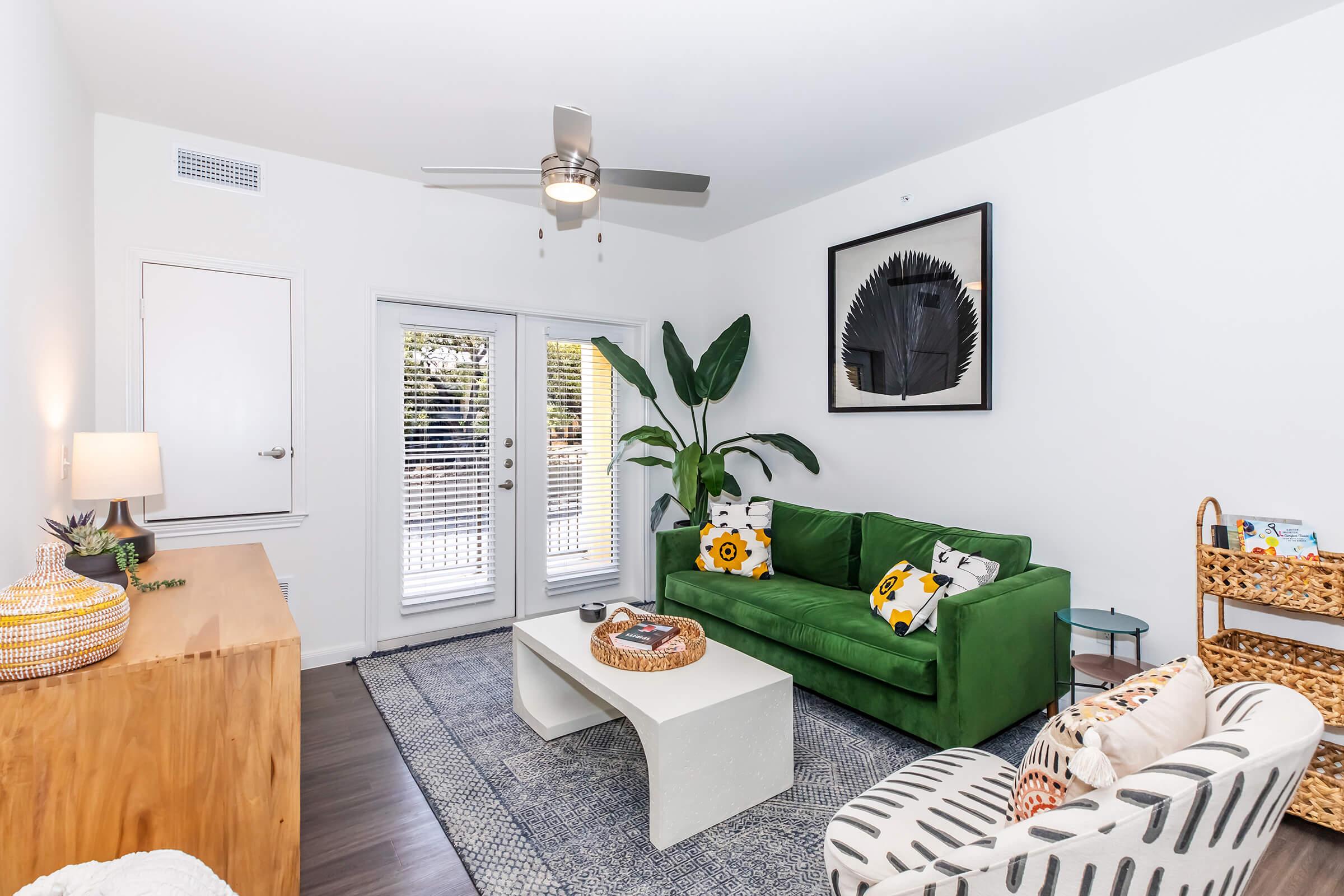 A modern living room featuring a green sofa adorned with patterned cushions, a white coffee table, and a stylish rug. Natural light filters in through large glass doors with blinds, revealing a view of the outdoors. A ceiling fan and potted plant add to the room's decor. A wooden sideboard and woven basket are visible.