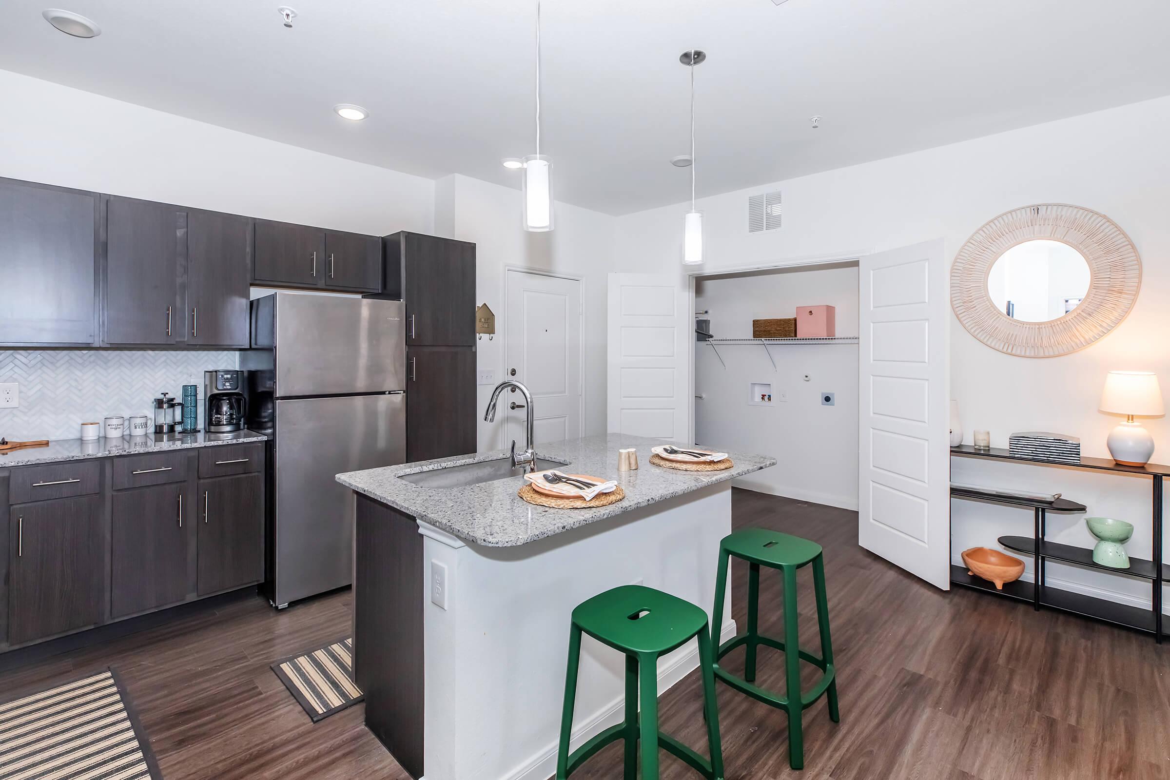 Modern kitchen with dark wooden cabinets, stainless steel refrigerator, and a gray granite island featuring green bar stools. There are organized shelves with decorative items and a round mirror on the wall, creating a stylish, contemporary living space. Natural light brightens the room.