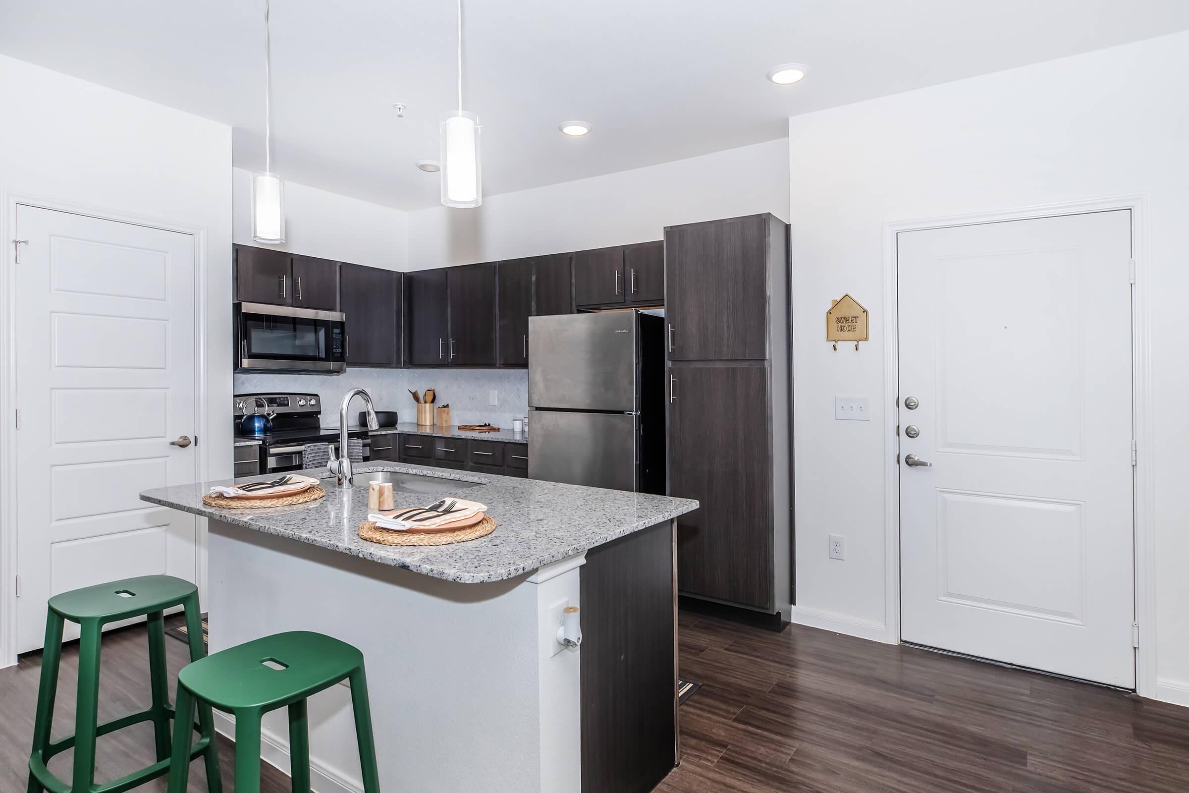 Modern kitchen featuring a gray granite island with two green stools, dark wood cabinets, stainless steel appliances including an oven and refrigerator, and a contemporary design. Bright lighting and a front door are visible, creating an inviting atmosphere.