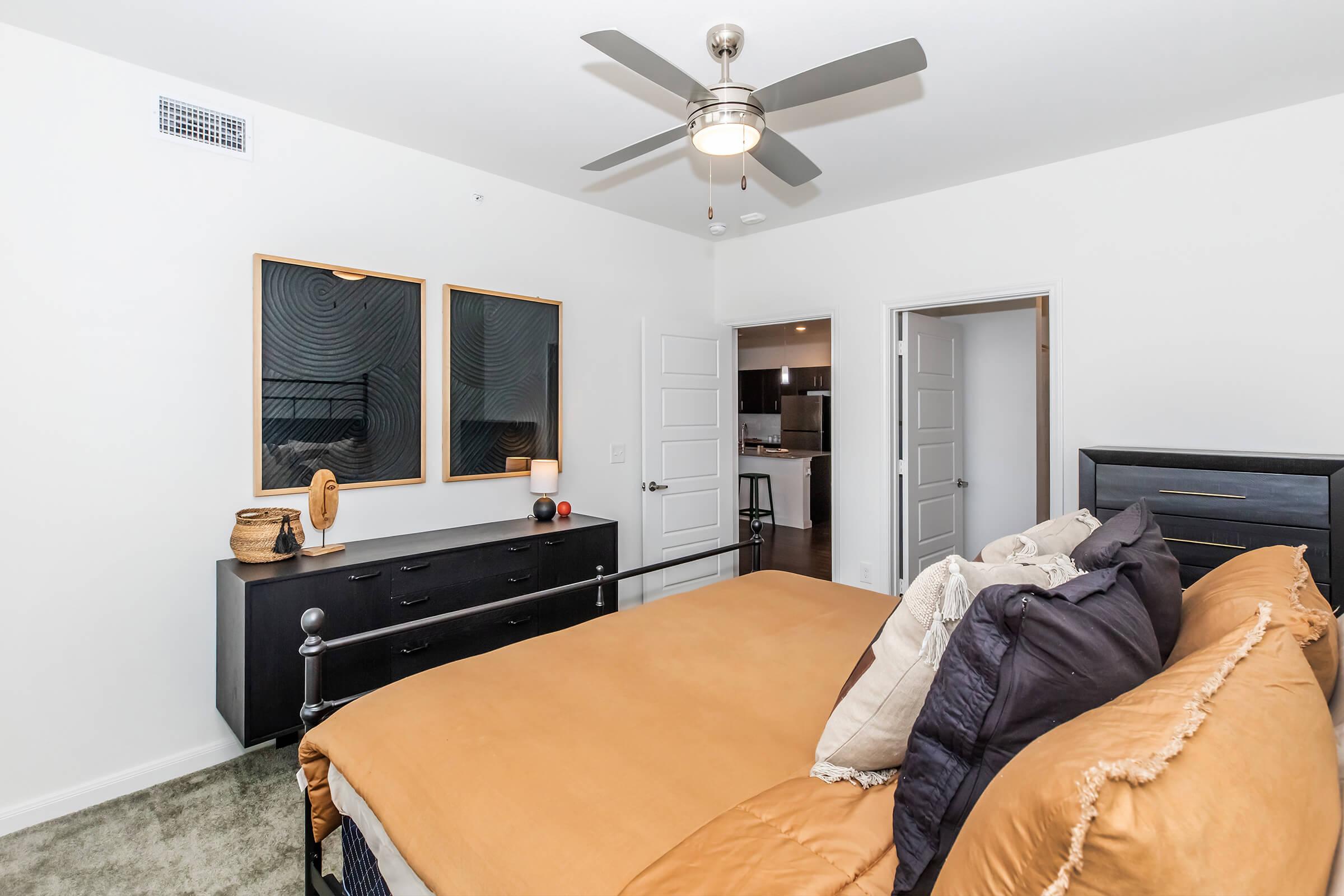 A modern bedroom featuring a large, inviting bed with an orange-brown duvet. Two framed abstract artworks hang above a sleek black dresser with a lamp and decorative items. A ceiling fan provides airflow, and two doors lead to other areas, giving the room a spacious feel with neutral-colored walls.