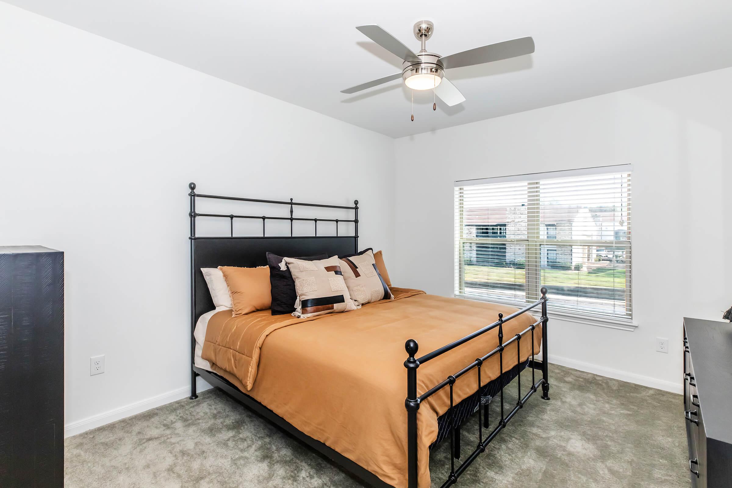 A cozy bedroom featuring a dark metal bed frame with an orange and beige bedding set. There is a decorative pillow on the bed, a ceiling fan above, and a window with white blinds allowing natural light to fill the room. The walls are painted white, and the floor is covered with light green carpet.