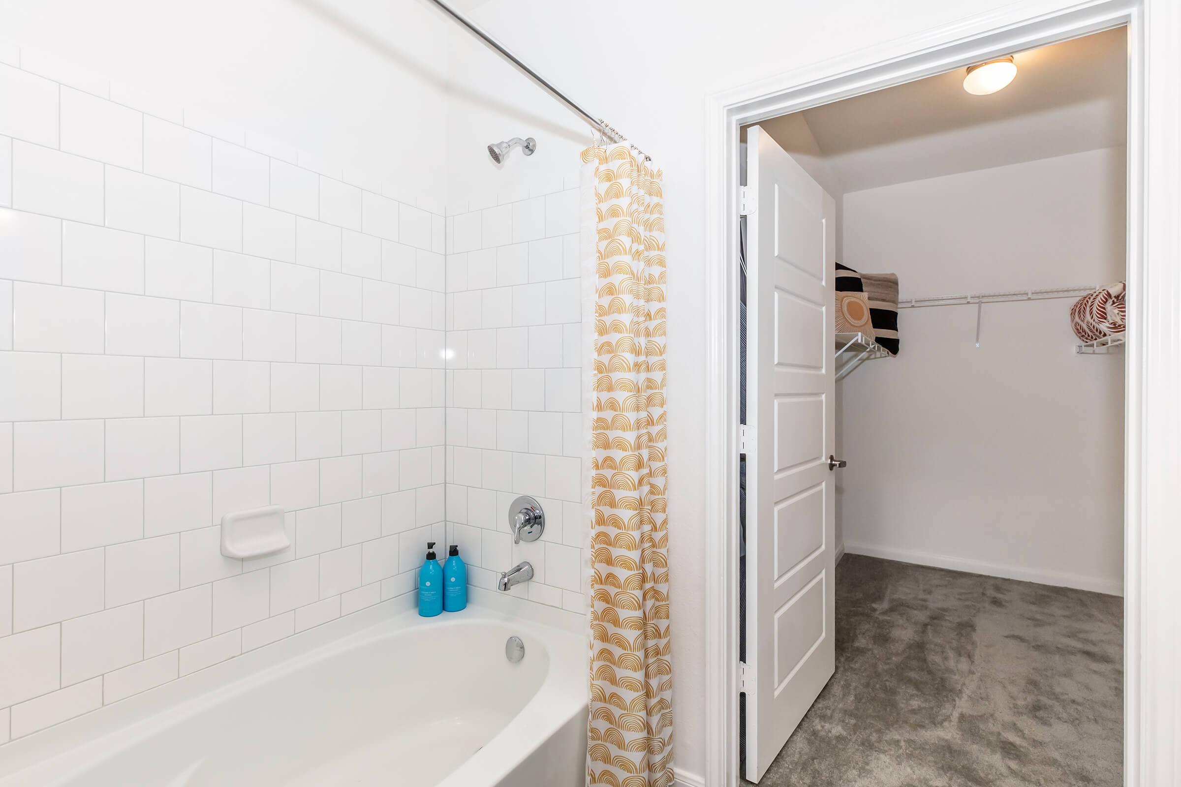 Bathroom featuring a bathtub with a shower curtain, white tiled walls, and two blue shampoo bottles on the edge of the tub. A door leads to a walk-in closet with carpeted flooring, and there is a ceiling light in the corner providing illumination.