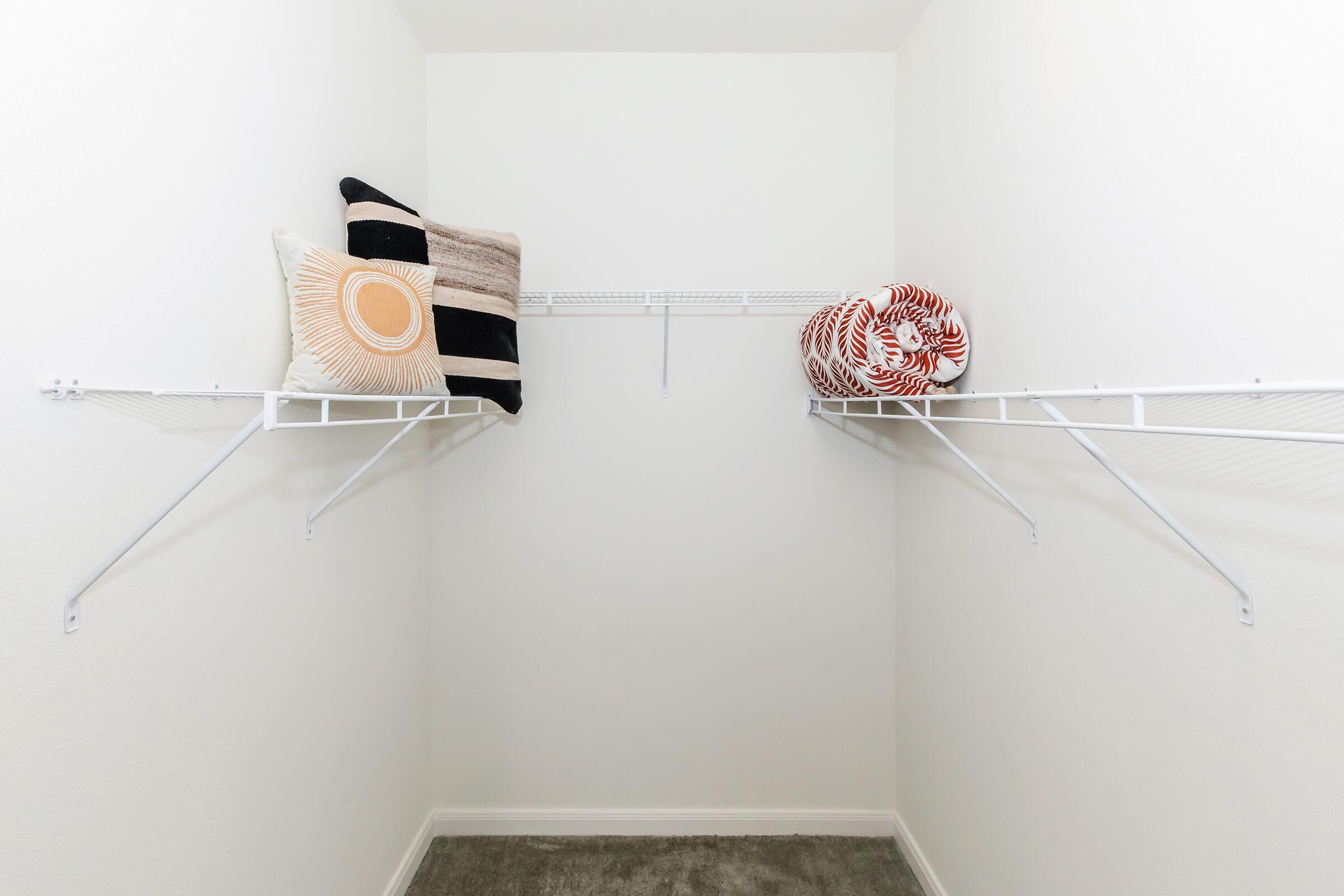 A clean, empty closet with two white wire shelves. On the shelves are two decorative pillows, one with a sunburst design and another with horizontal stripes. The walls are white, and the carpeted floor is a light gray color.