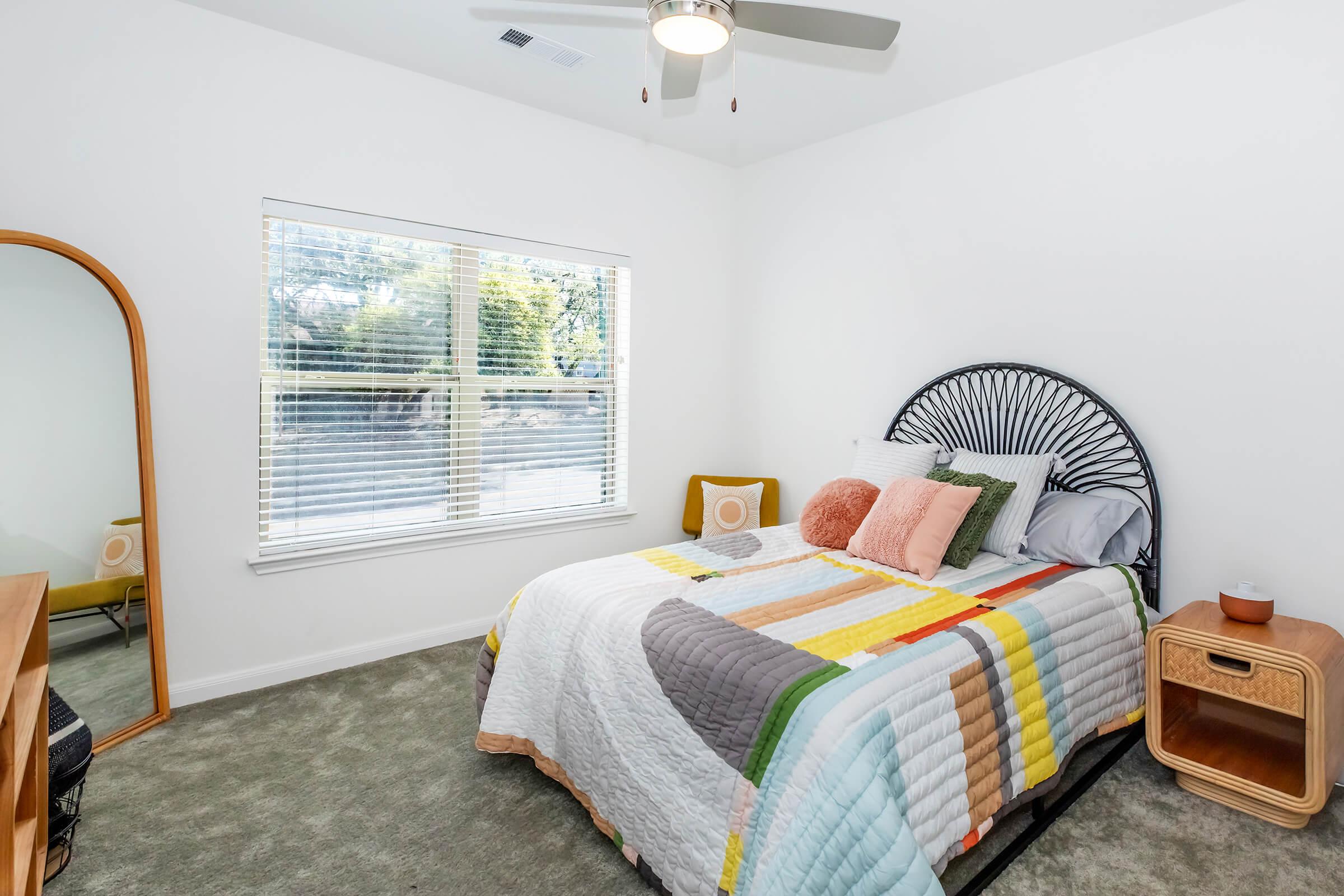 A well-decorated bedroom featuring a bed with colorful bedding, a large window with natural light, a round mirror, a stylish side table, and a cozy carpet. The room has a modern aesthetic with a mix of textures and soft colors, creating a warm and inviting atmosphere.