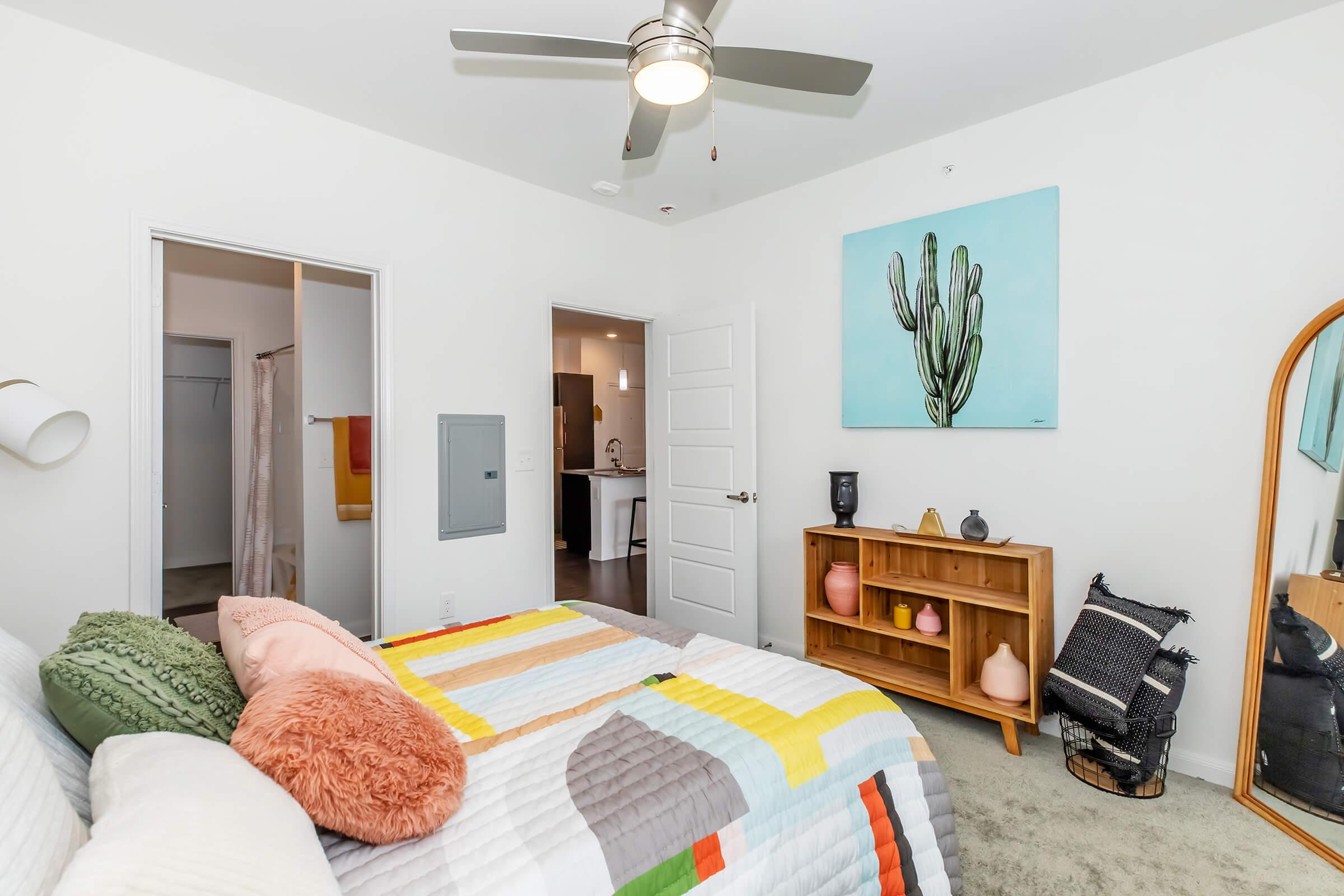 A brightly lit bedroom featuring a bed with a colorful quilt, a cactus artwork on the wall, and a wooden shelving unit with decorative items. A ceiling fan is installed above, and a large mirror is positioned against the wall. The room has a warm and inviting atmosphere.