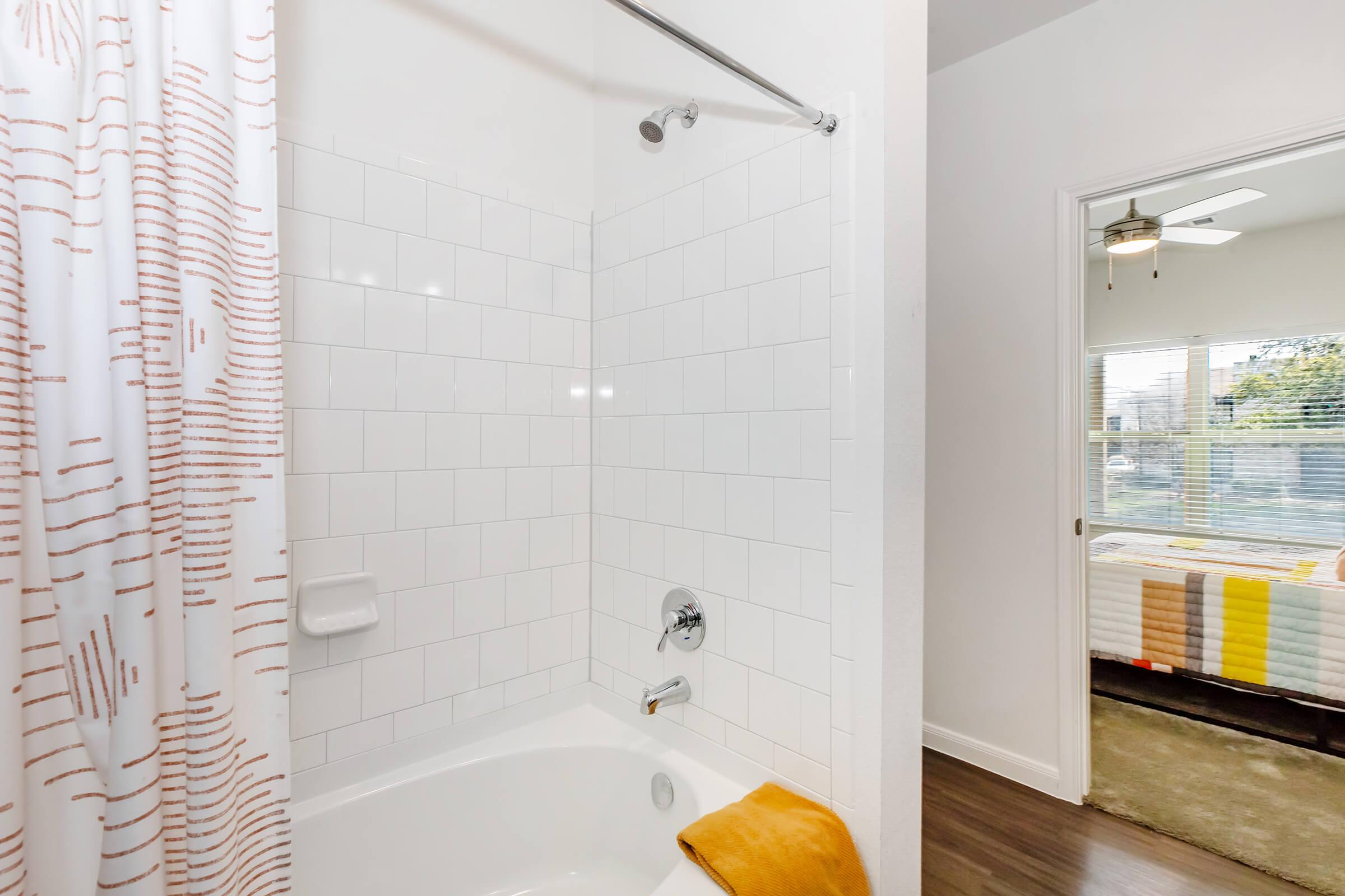 A bright and modern bathroom featuring a white tiled bathtub with a chrome showerhead. The shower curtain has a simple design, and a plush yellow towel is draped over the tub. A doorway leads to another room with a window demonstrating natural light, and a glimpse of colorful bedding is visible.