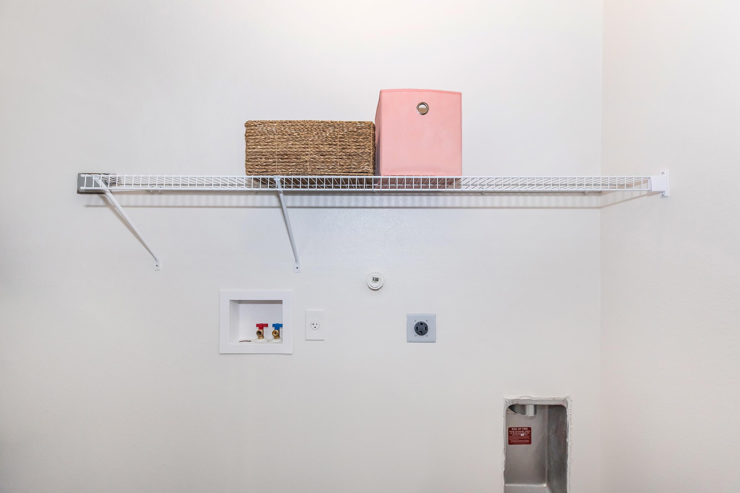 A white wire shelf mounted on a light-colored wall, holding a pink container and a woven basket. Below the shelf, there’s a small recessed box with colorful buttons and a power outlet. A small hole in the wall is visible on the right side.