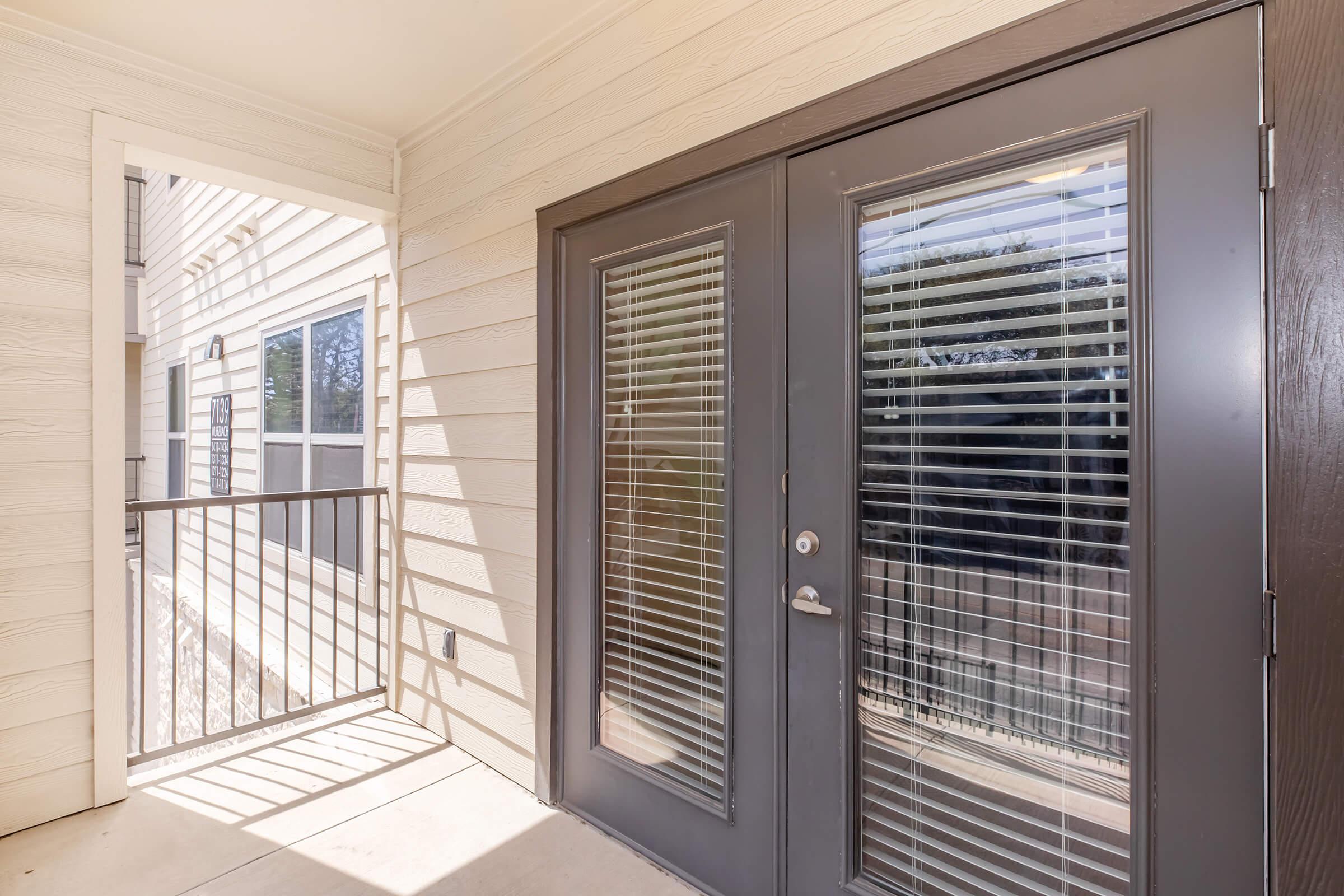 A doorway with double glass doors featuring horizontal blinds, leading to a small balcony area. The exterior walls are light-colored, with a railing visible on one side. Natural light illuminates the space, highlighting a clean and modern design.