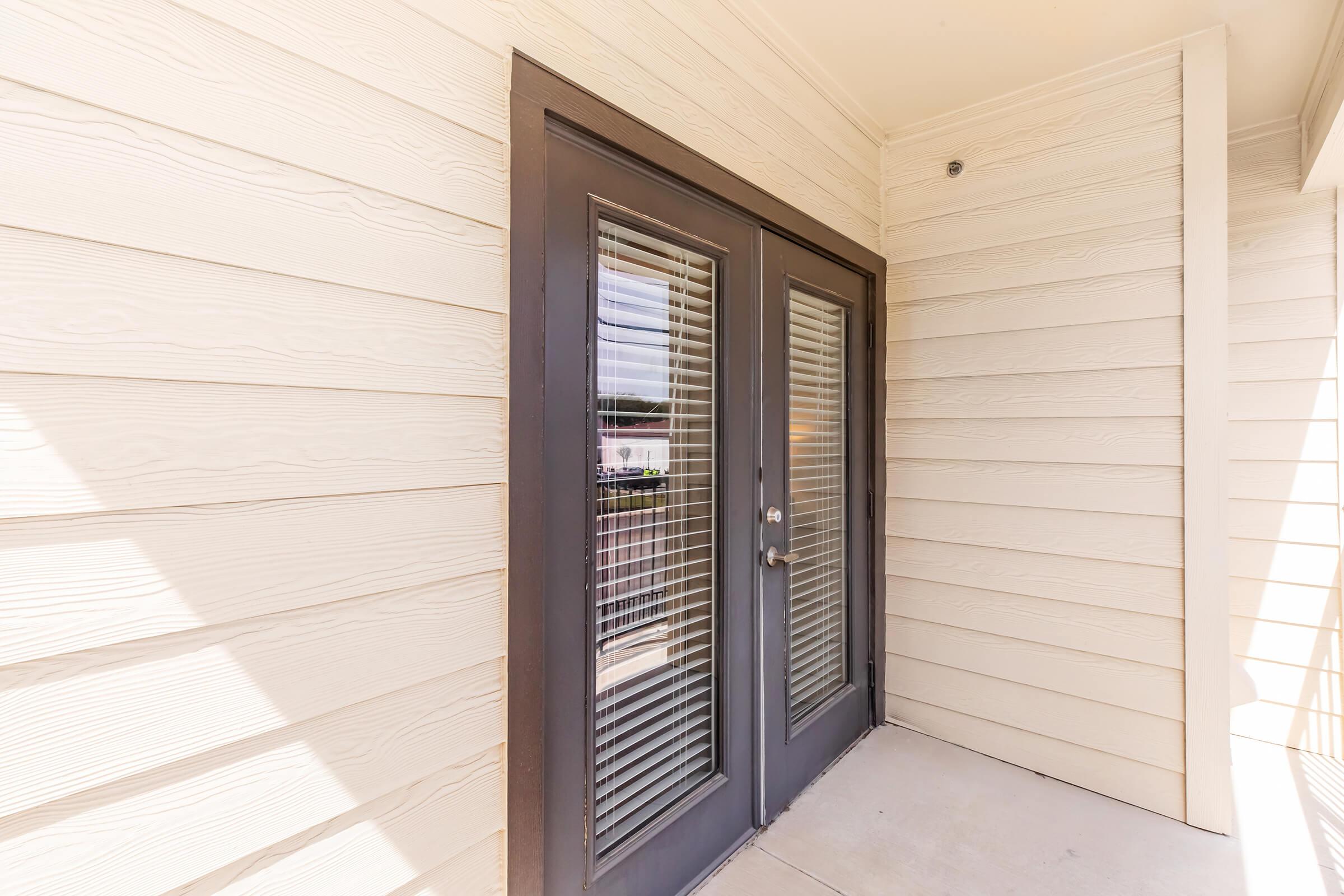 Double glass doors with horizontal blinds, framed by a beige exterior wall. The entrance area is well-lit, showcasing a clean and modern design.
