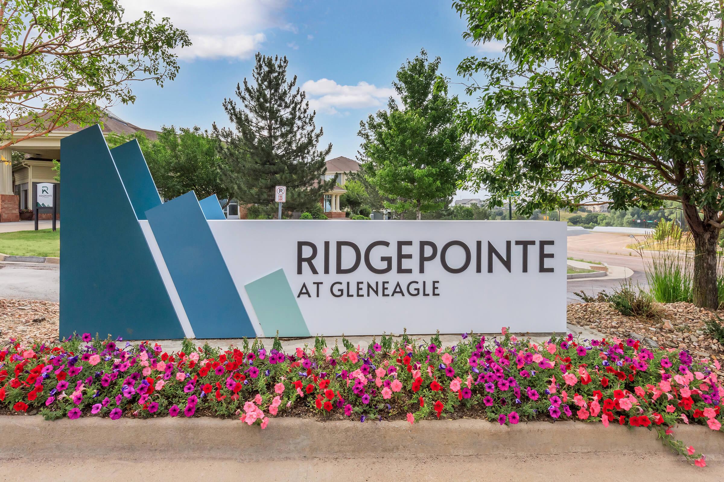 Sign for Ridgepointe at Gleneagle, featuring a modern design with geometric shapes in shades of blue and green. The sign is surrounded by colorful flower beds and greenery, set against a backdrop of buildings and a clear sky.
