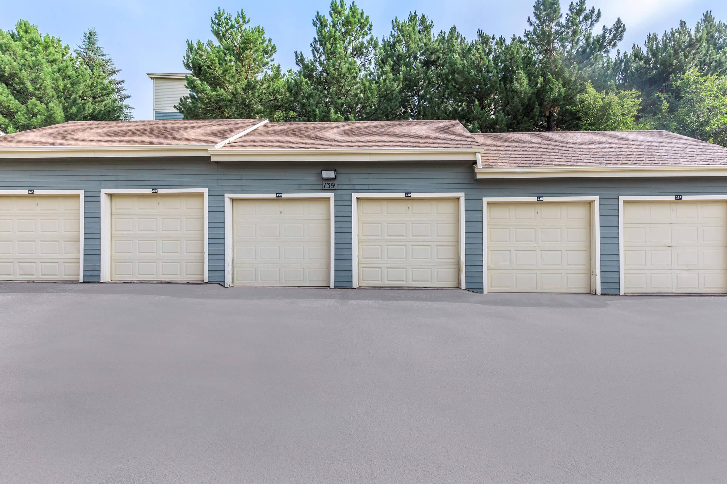 Row of five garage doors in a residential area, featuring beige doors with windows, set against a backdrop of green trees and a blue house exterior. The garages are lined up symmetrically on a paved surface, with clear skies above, creating a neat and orderly appearance.