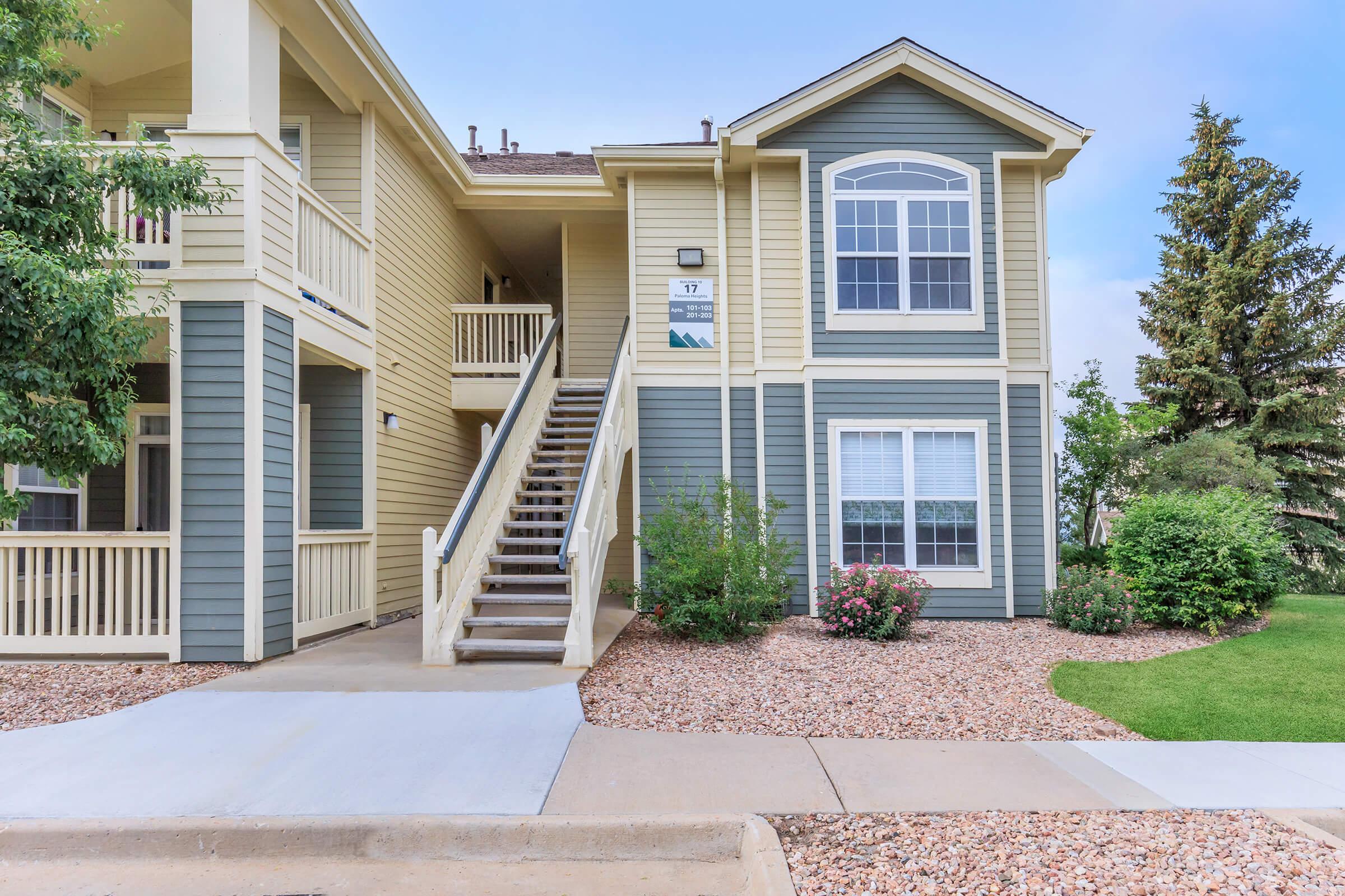 Two-story apartment building with beige and blue siding. A set of stairs leads up to a second-floor entrance, featuring a large window. The surrounding area includes well-kept shrubs and grass, with a sidewalk and gravel landscaping visible in front. Clear blue sky overhead.