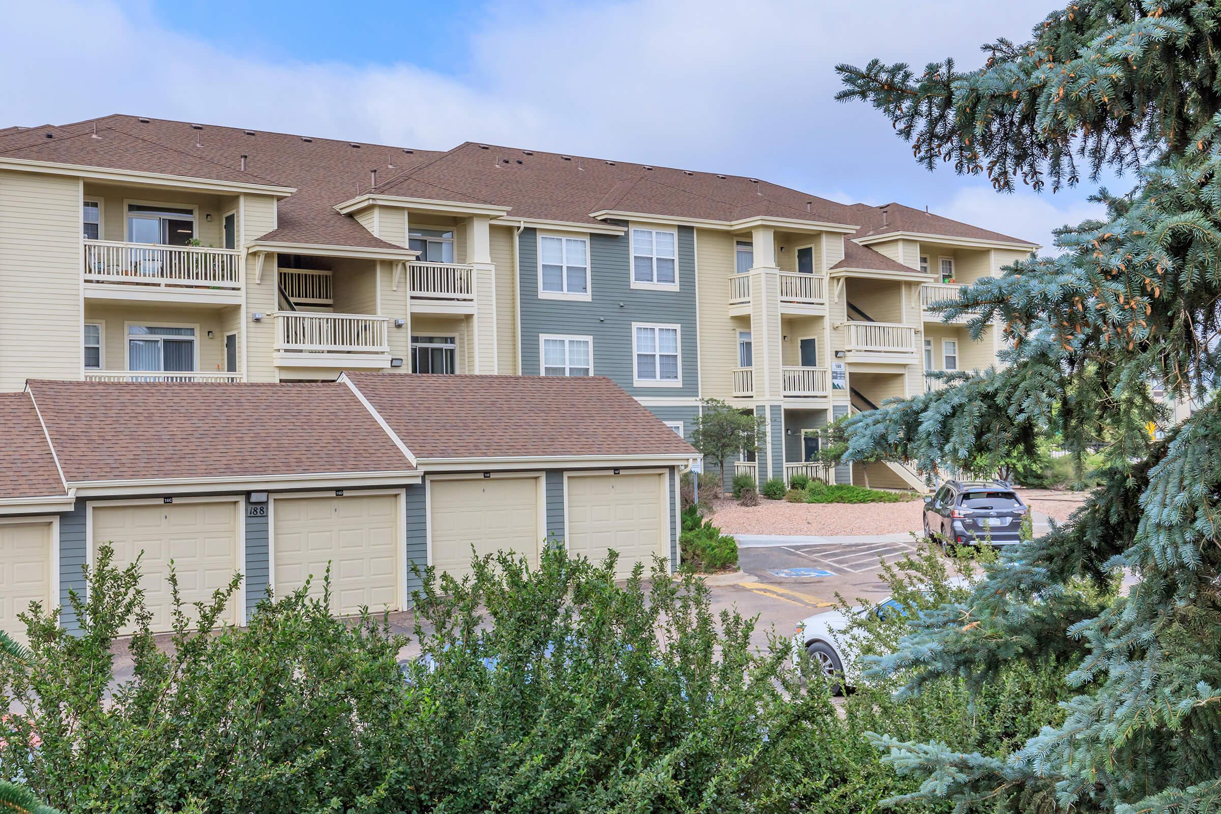 A multi-story apartment building with balconies, surrounded by greenery. In the foreground, there are garage units, and in the background, cars are parked. The sky is partly cloudy, providing a bright and welcoming atmosphere.
