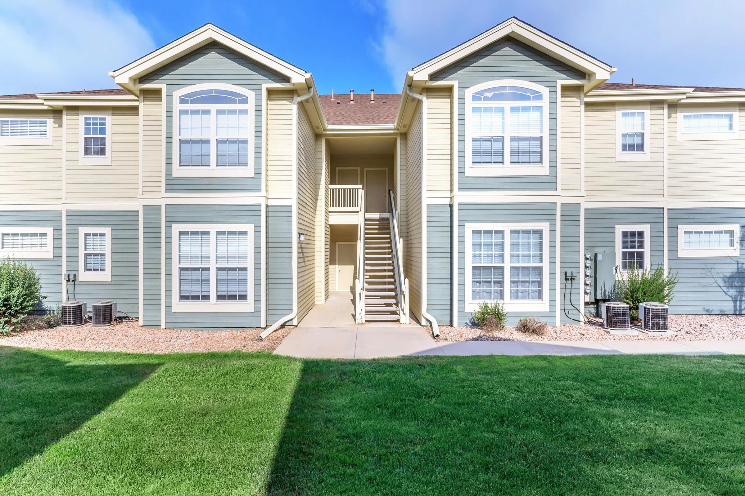 A multi-unit residential building featuring two levels with beige and blue siding, large windows, and a central staircase. A well-maintained lawn and shrubs surround the entrance, with air conditioning units visible on the ground. The sky is partly cloudy, creating a bright atmosphere.