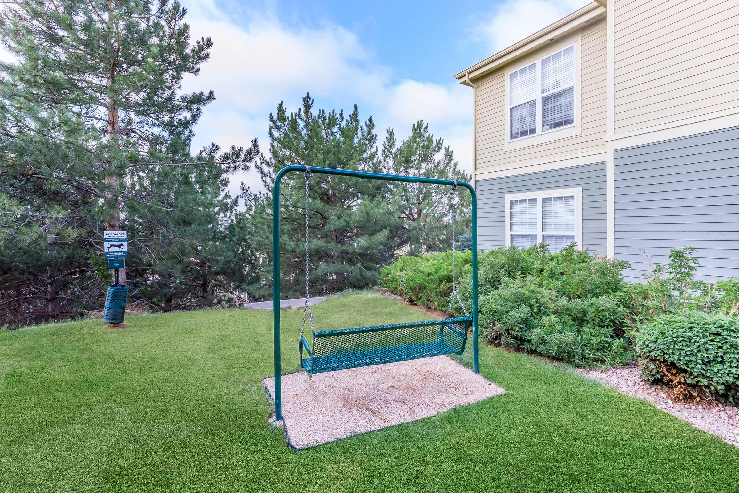 A green swing set on a grassy area surrounded by trees, with a gravel base for stability. In the background, a multi-story building features beige and gray siding. The sky is partly cloudy, creating a serene outdoor setting ideal for relaxation.
