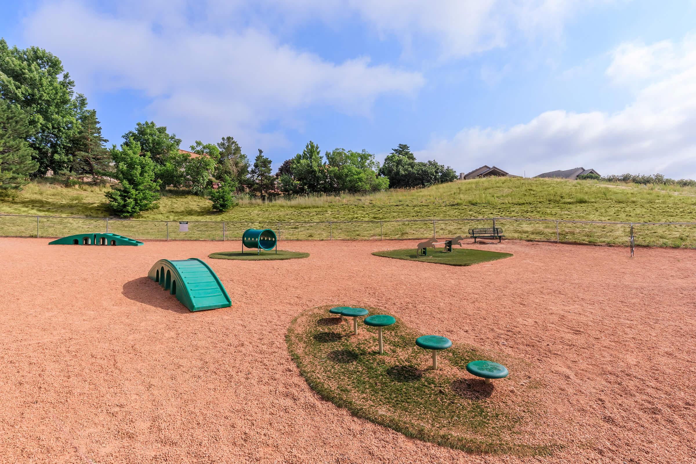 A playground featuring a soft, sandy surface with various green equipment, including climbing structures and benches. Trees and grassy hills in the background under a partly cloudy sky, creating a tranquil outdoor setting for play and recreation.