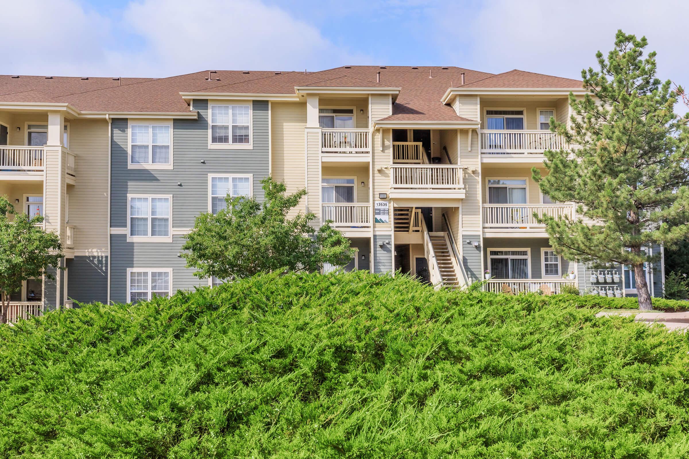 Three-story apartment building with beige and gray siding, featuring balconies and surrounded by greenery, including a low bush in the foreground. The sky is partly cloudy, and there are trees nearby. A staircase leads to the entrance of the building.