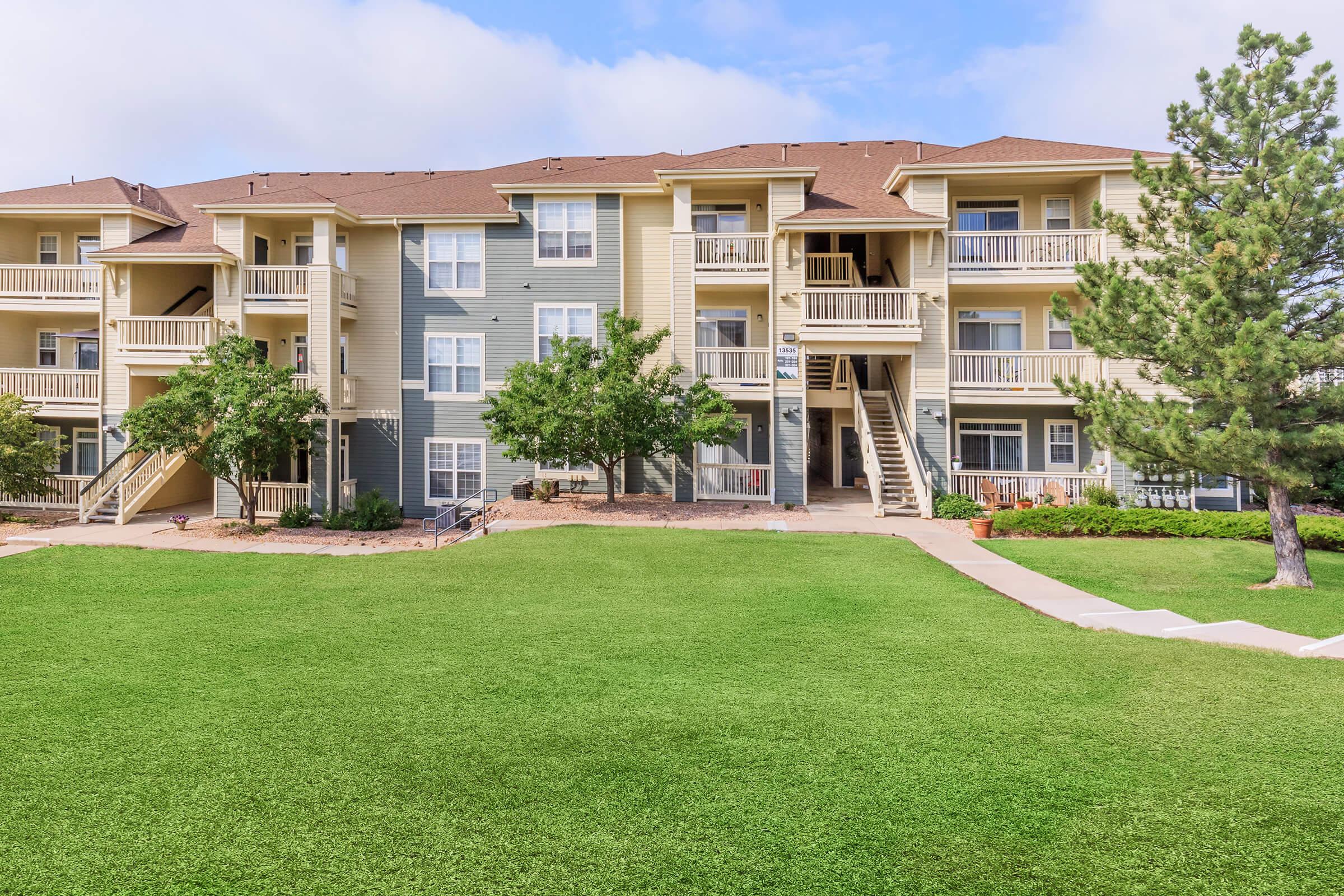 A multi-story apartment building featuring balconies, surrounded by well-maintained green grass and trees. A paved walkway leads to the entrance, with blue skies and fluffy clouds in the background. The overall setting is bright and inviting, showcasing a residential community.