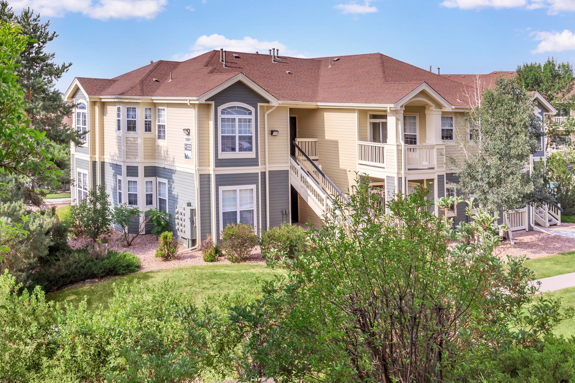 A view of a multi-story apartment building surrounded by lush greenery and landscaping. The building features a combination of yellow and blue siding, with balconies and large windows. A well-maintained lawn and trees are visible in the foreground, contributing to a peaceful residential atmosphere.