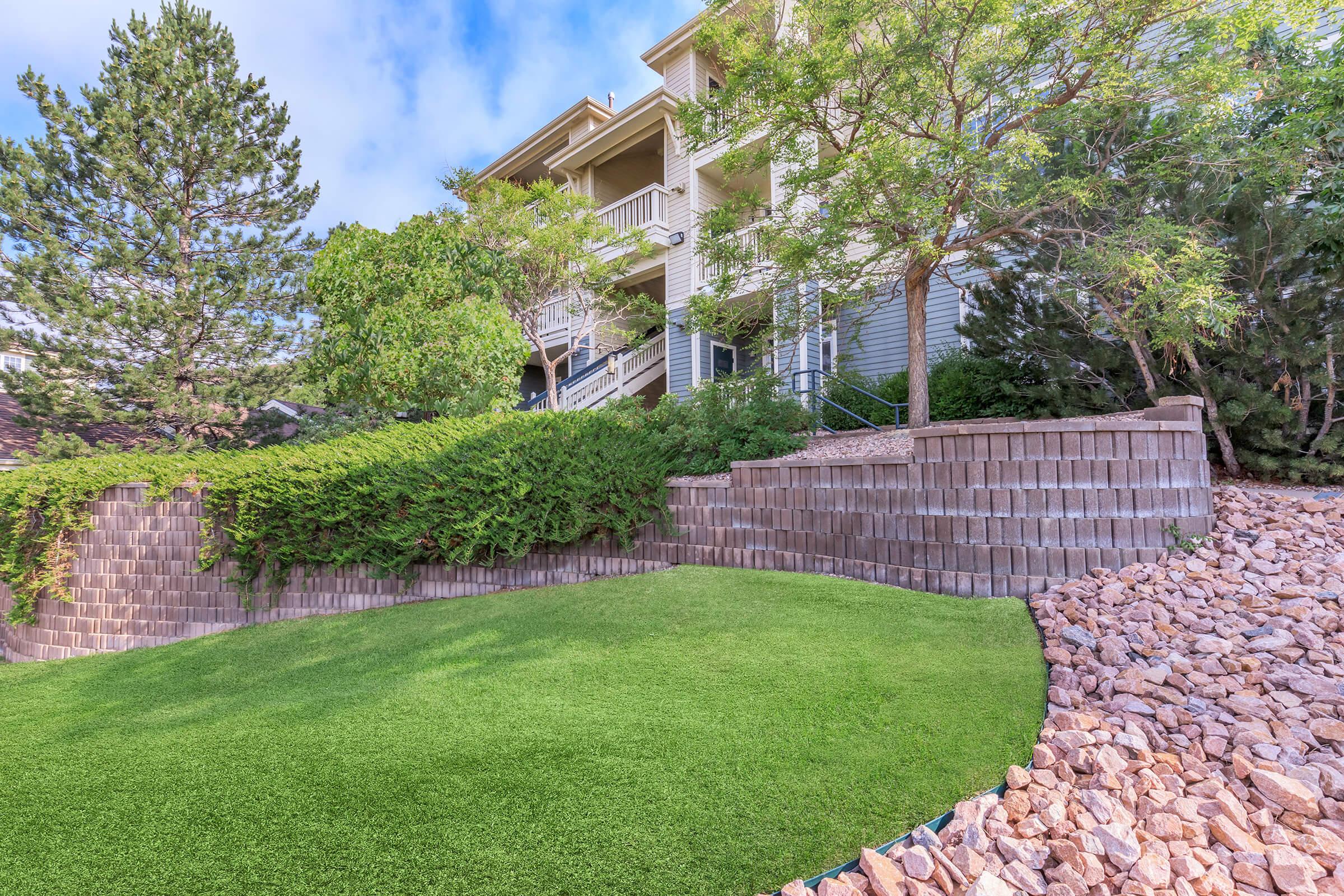 A well-maintained landscape featuring a lush green lawn bordered by neatly arranged rocks and a low stone wall. In the background, a multi-level building is partially visible, surrounded by trees and shrubs under a clear blue sky.