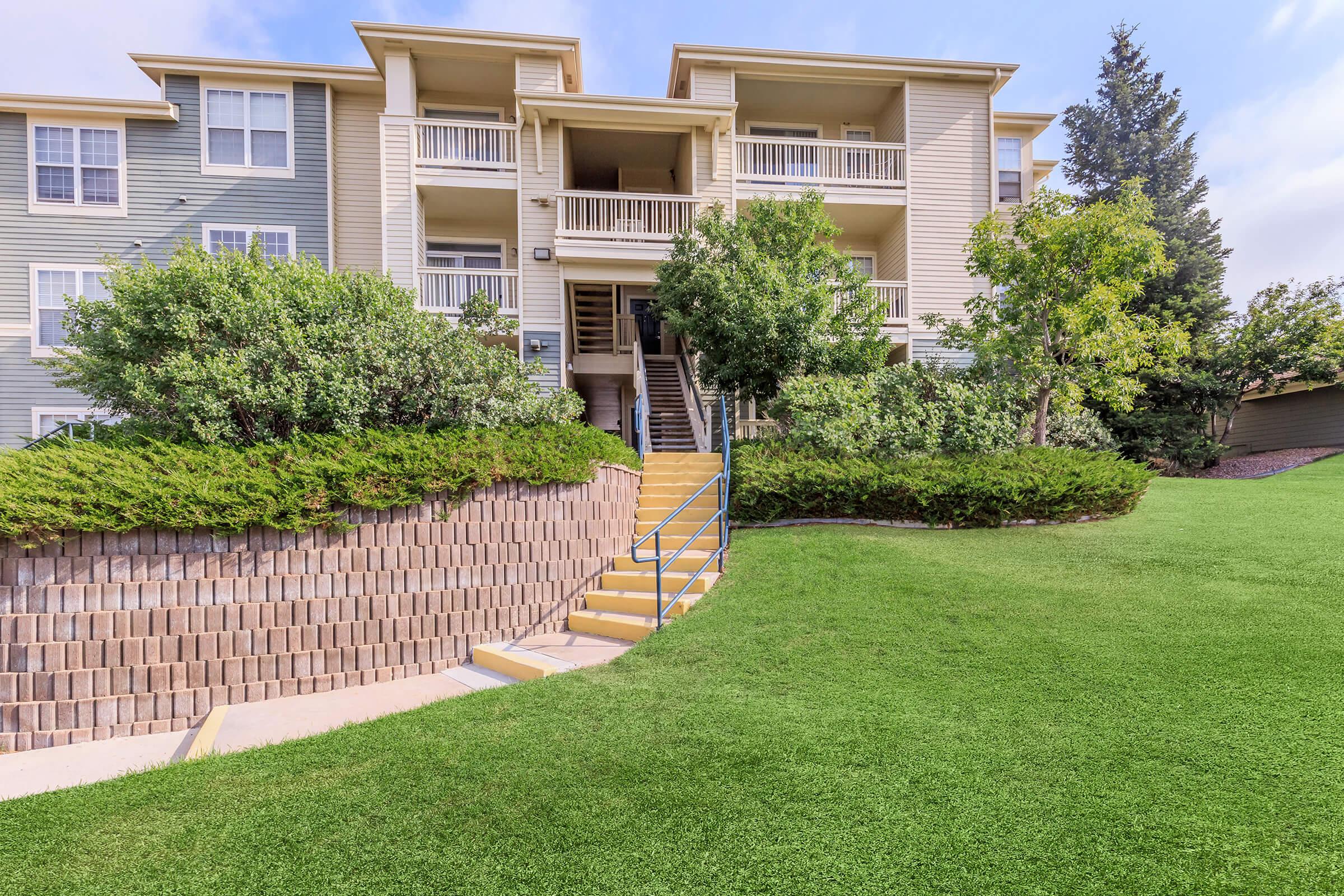 View of a three-story apartment building surrounded by neatly landscaped greenery. A set of stairs leads up from a grassy area to the entrance, flanked by bushes and trees, creating a welcoming outdoor space. The architecture features light-colored siding with balconies on the upper floors.
