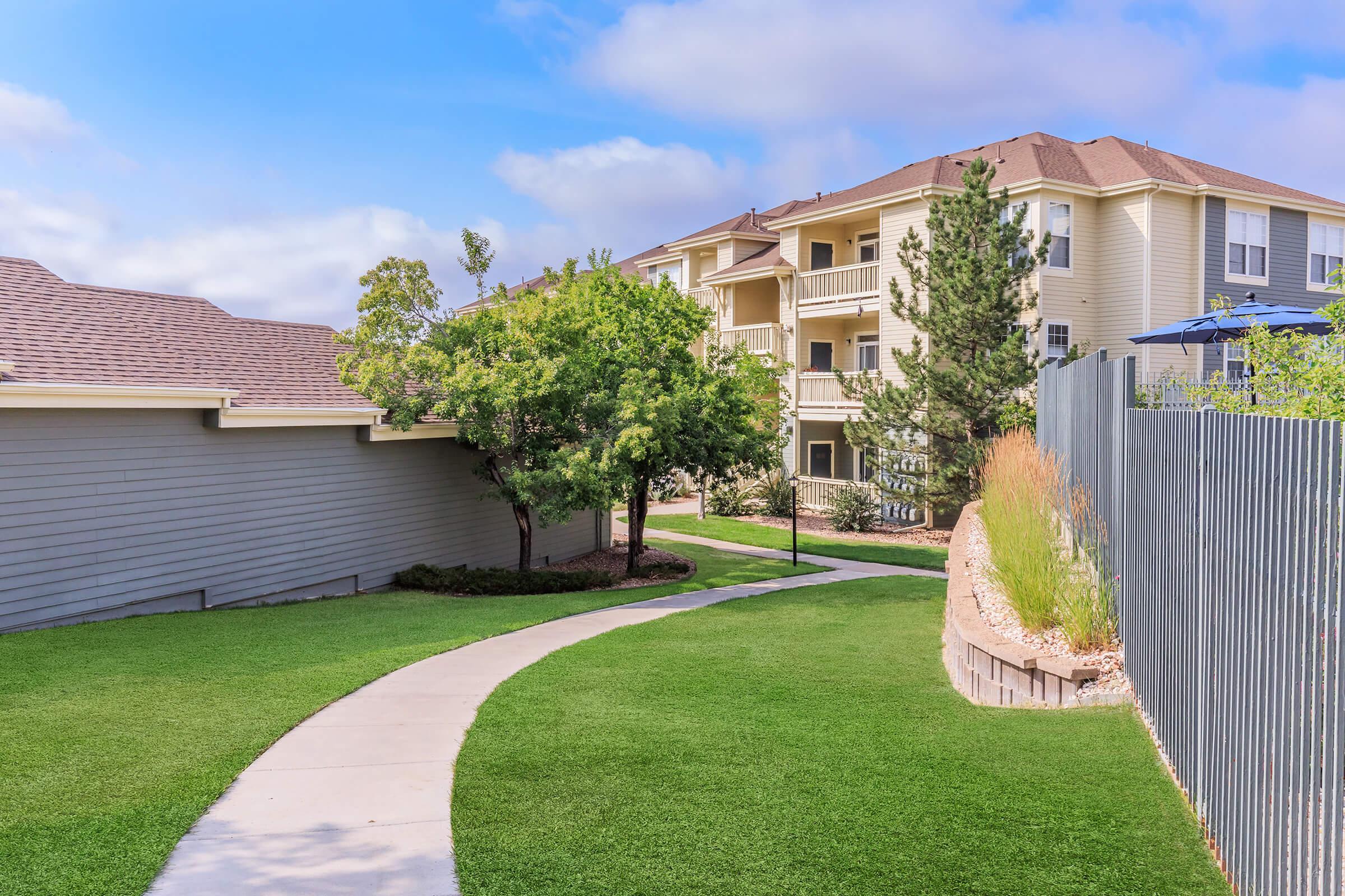 A serene landscape featuring a curved pathway lined with green grass, leading past a modern multi-story apartment building. Trees and shrubs complement the picturesque scene, with a clear blue sky and soft clouds in the background. The setting conveys a peaceful residential atmosphere.