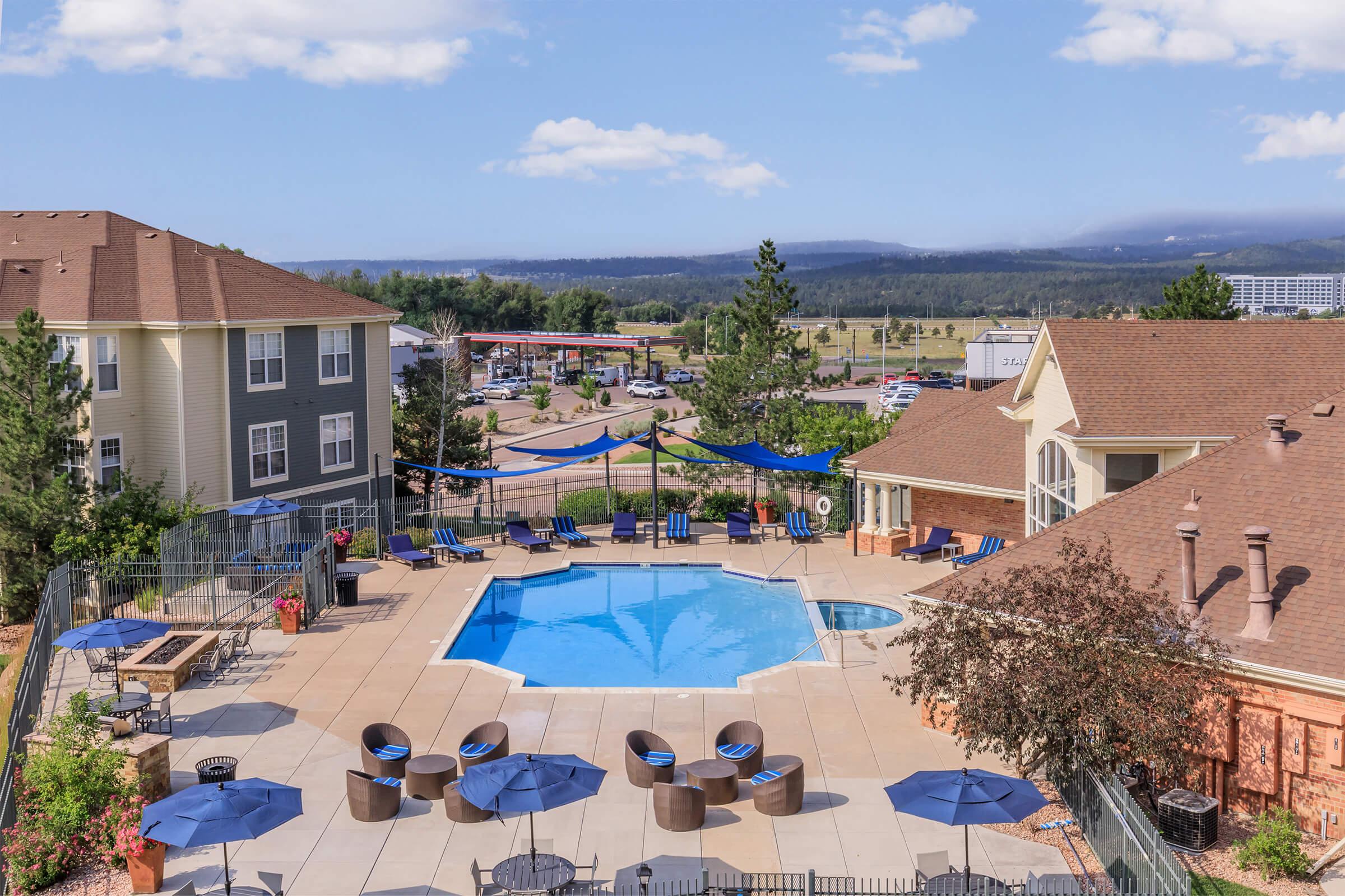 Aerial view of a hotel pool area featuring a rectangular swimming pool surrounded by lounge chairs and umbrellas. The landscape includes trees, blue skies, and distant mountains, with nearby buildings and a parking area visible in the background.