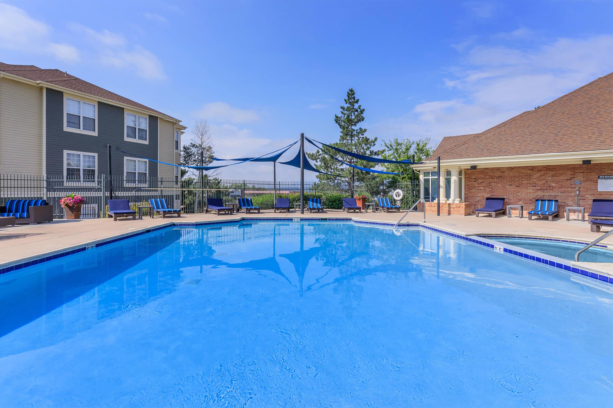 A clear swimming pool with blue water, surrounded by lounge chairs with blue and black cushions. In the background, there are two buildings and some trees under a partly sunny sky. A shaded area with a canopy is above the poolside seating, creating a relaxing atmosphere.