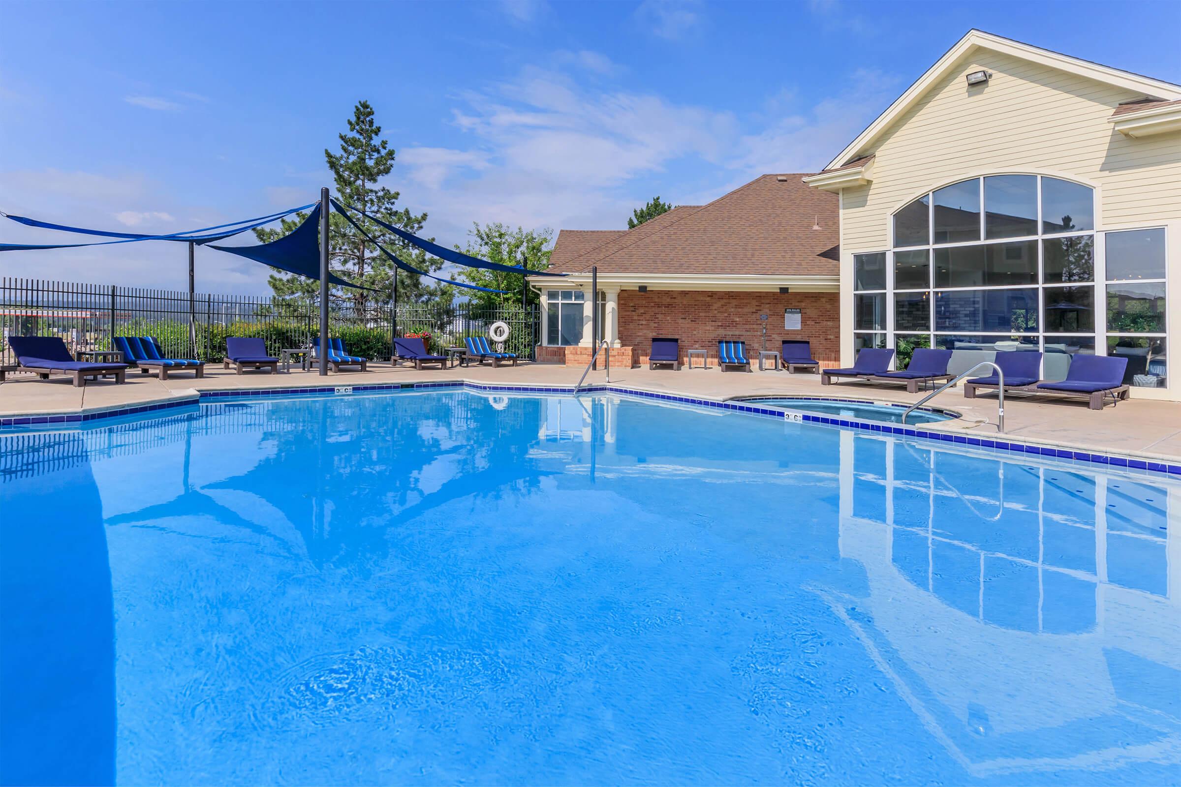 A clear blue swimming pool surrounded by lounge chairs and shaded areas. The pool area is well-lit, featuring a modern building with large windows in the background and a beautiful sky above. Ideal for relaxation and leisure.