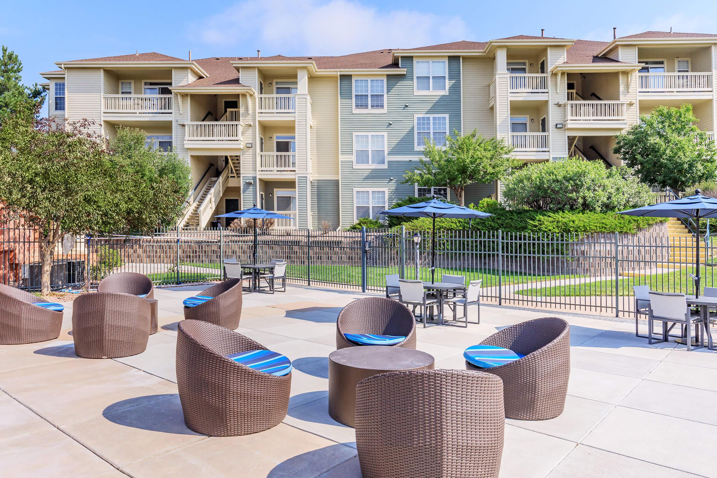 A spacious outdoor area featuring modern wicker chairs with blue cushions around small tables. In the background, there are multi-story residential buildings with balconies and well-maintained greenery. The setting suggests a pleasant atmosphere perfect for relaxation or socializing.