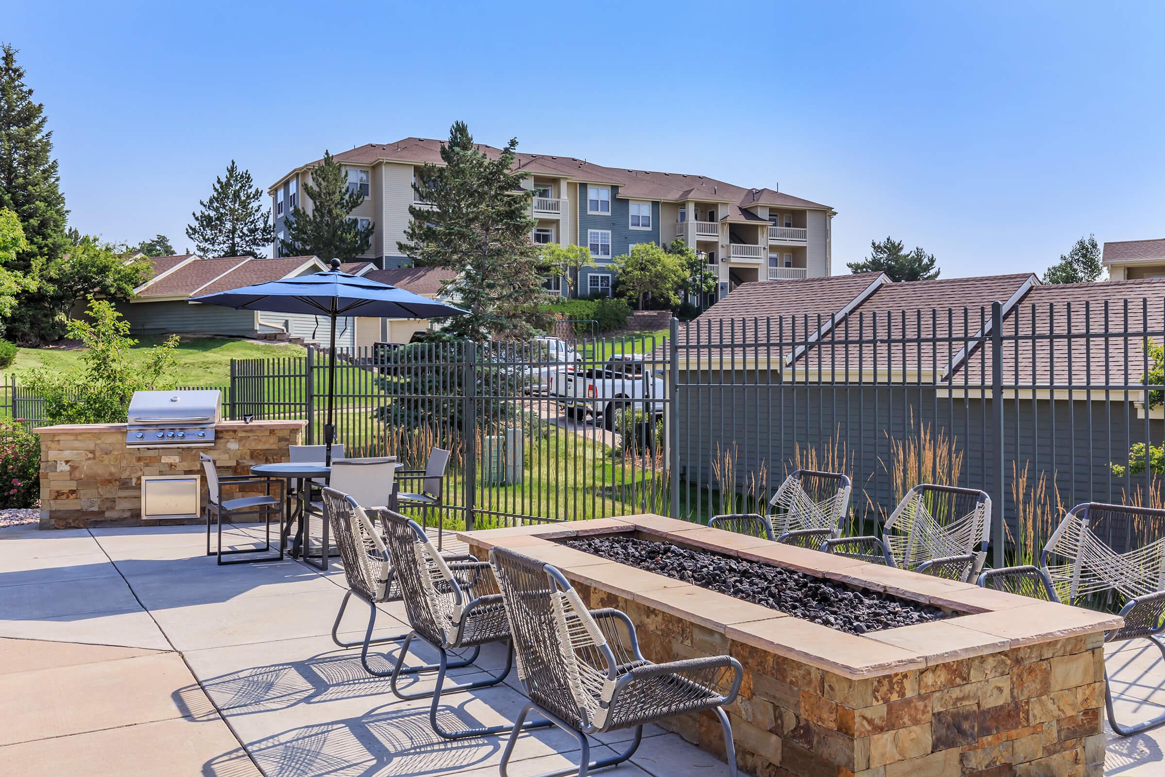 Outdoor patio area featuring a barbecue grill and a fire pit surrounded by chairs. In the background, there are multi-story apartment buildings and lush greenery under a clear blue sky. A fence encloses the space, providing a cozy setting for relaxation or gatherings.