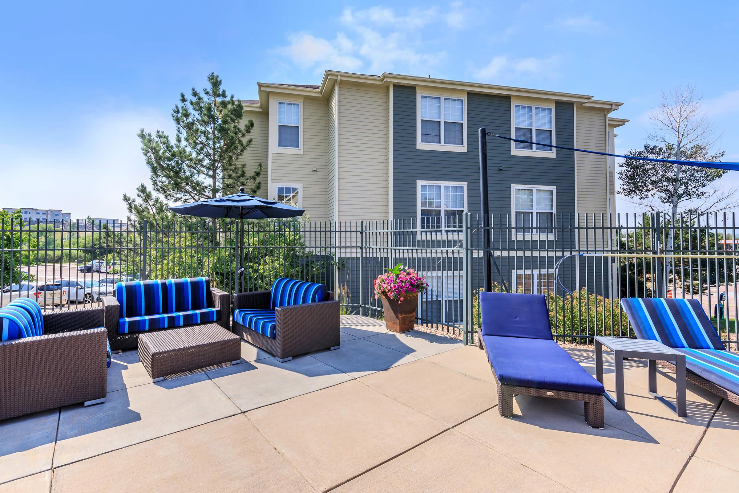 Outdoor lounge area featuring stylish seating with blue and striped cushions, a large umbrella, and potted flowers. In the background, a multi-story building is visible, surrounded by a fence. The scene is bright and inviting, perfect for relaxation.