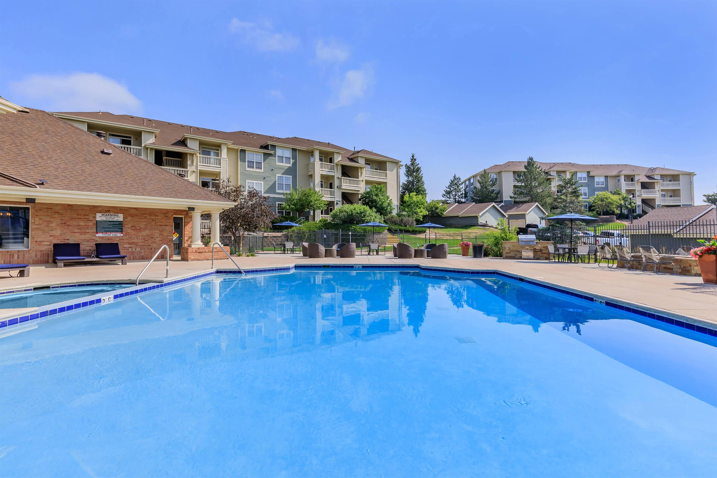 A bright blue swimming pool surrounded by well-maintained landscaping and lounge chairs. In the background, multiple residential buildings are visible against a clear blue sky. The scene suggests a relaxing outdoor area for residents or guests.