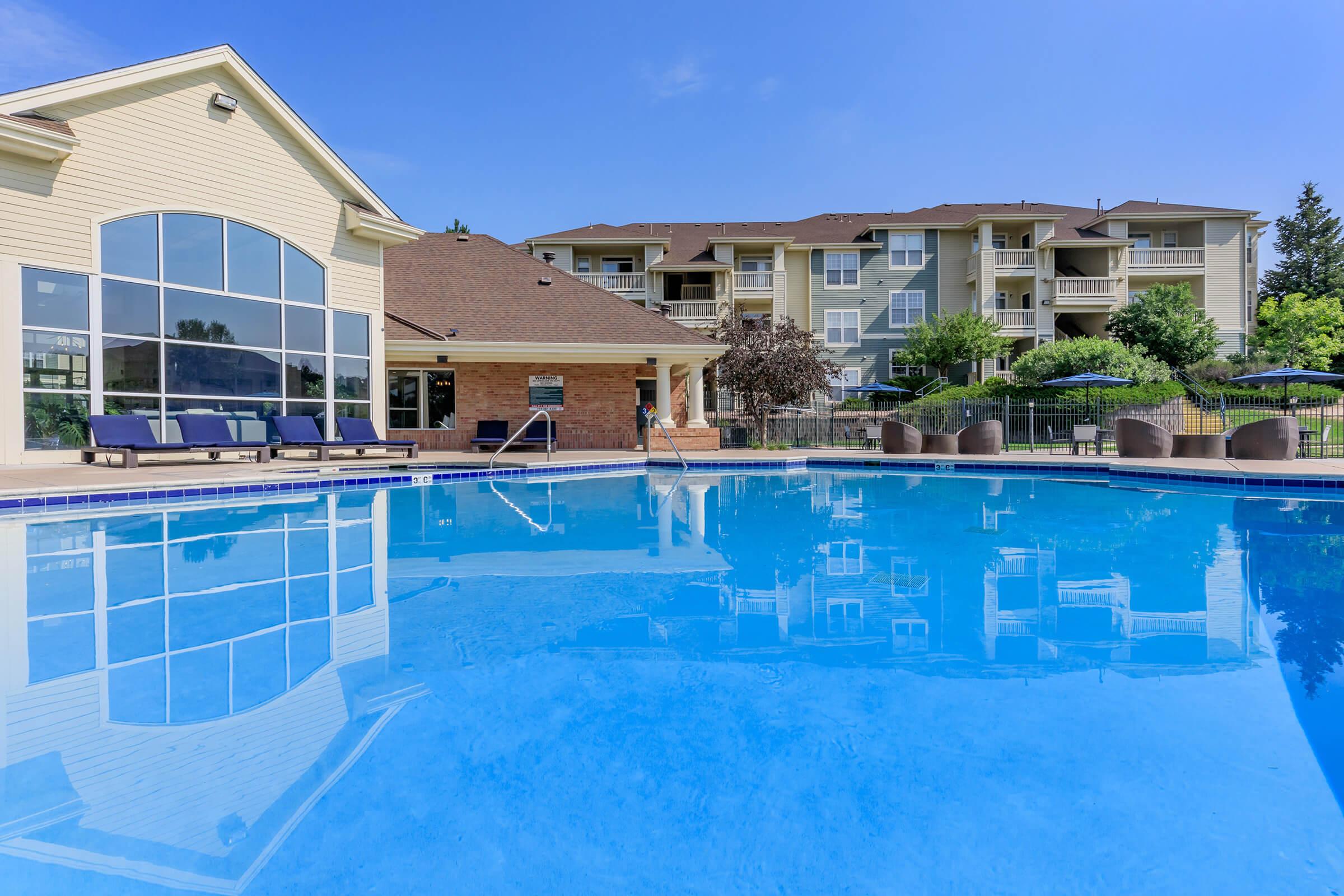 A clear blue swimming pool surrounded by lounge chairs and a building with large windows. In the background, there are residential apartments and trees, under a bright blue sky. The scene suggests a relaxing outdoor environment.