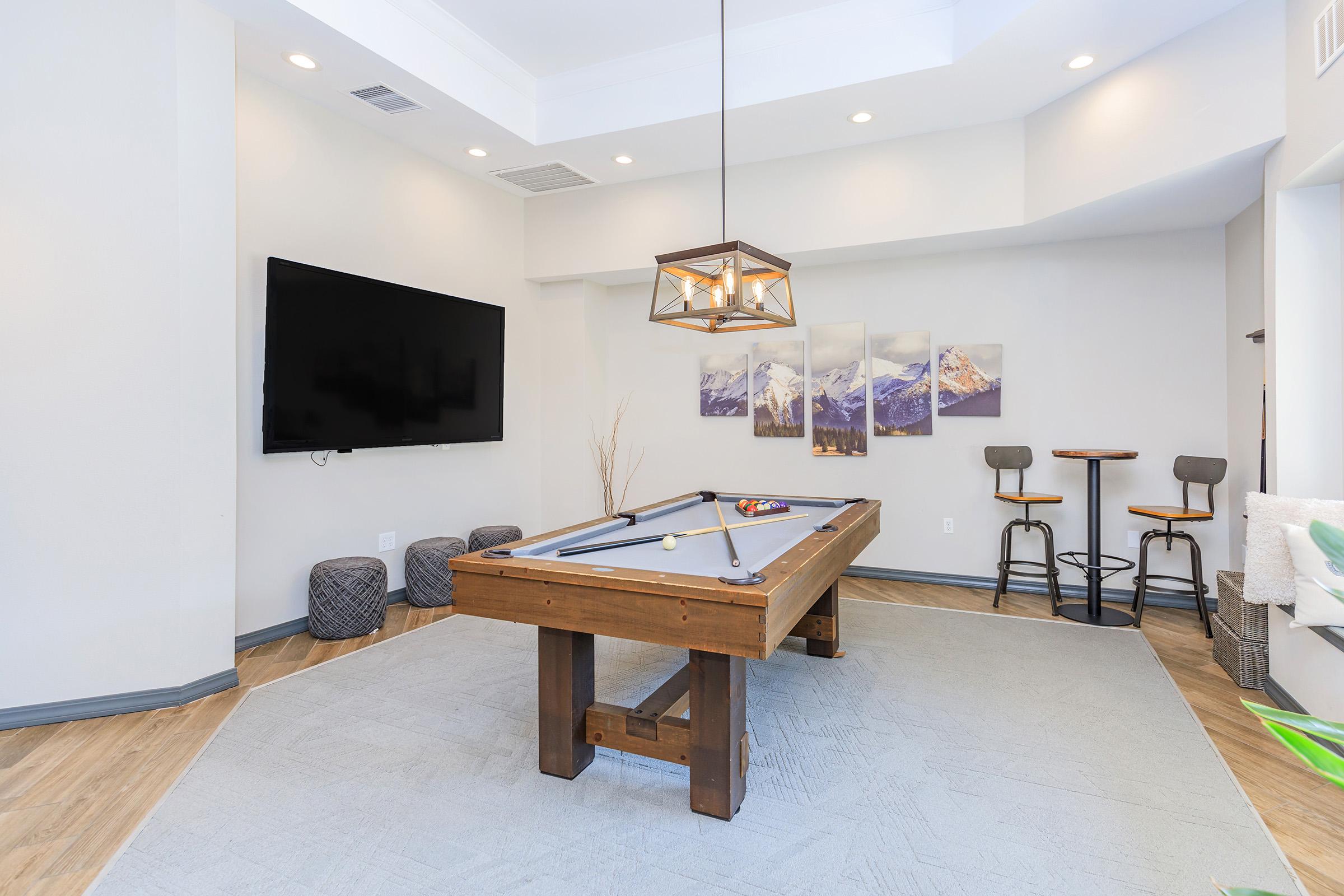 A modern game room featuring a sleek billiard table, a flat-screen TV mounted on the wall, and a stylish light fixture. The space includes cozy seating options and a decorative mountain landscape artwork. The design combines contemporary elements with a warm, inviting atmosphere.