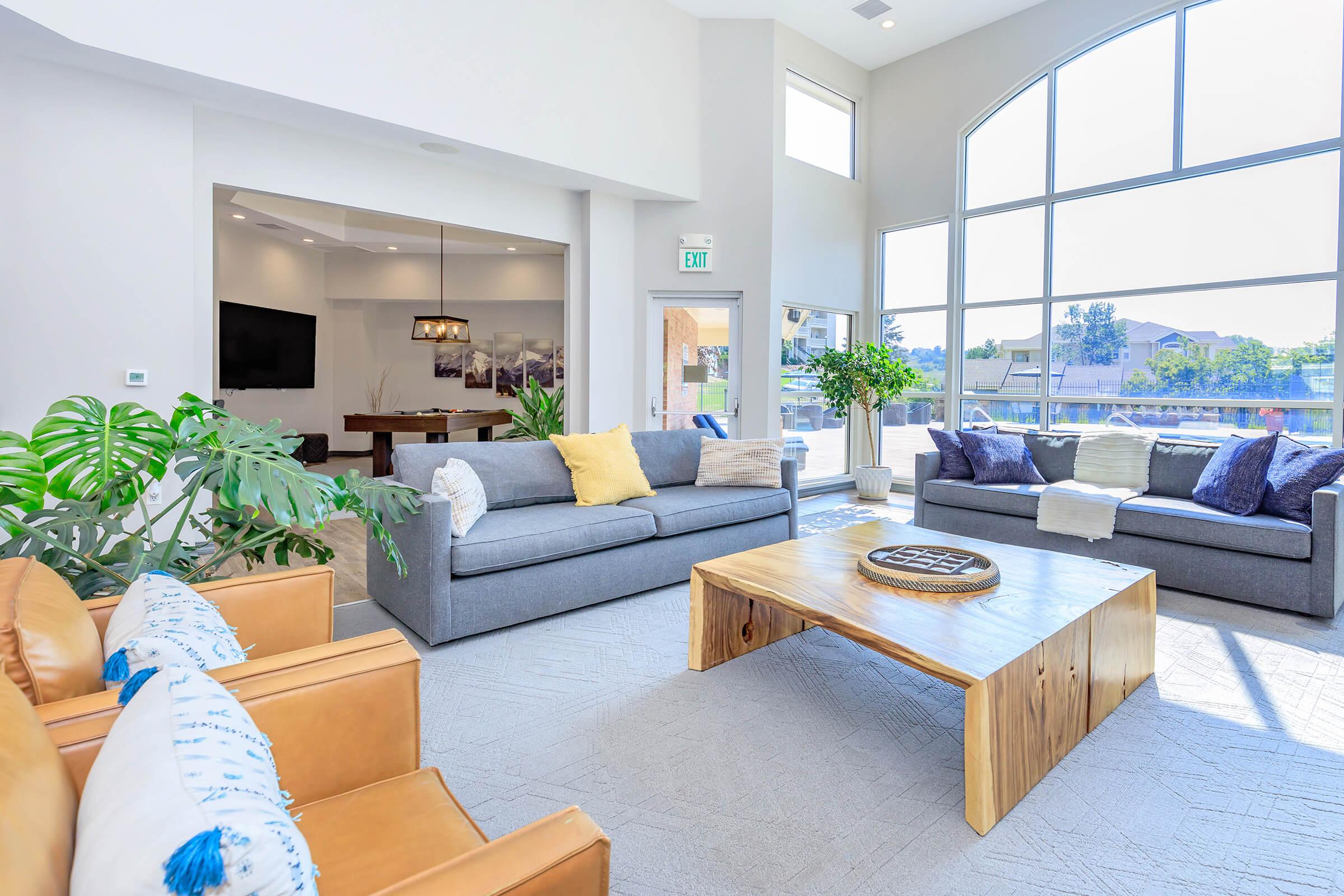 A modern living room featuring two gray sofas, a wooden coffee table, and brown leather armchairs. Large windows allow natural light to fill the space, revealing a view of the outdoors. Decorative pillows are scattered on the sofas, and potted plants add a touch of greenery to the inviting atmosphere.